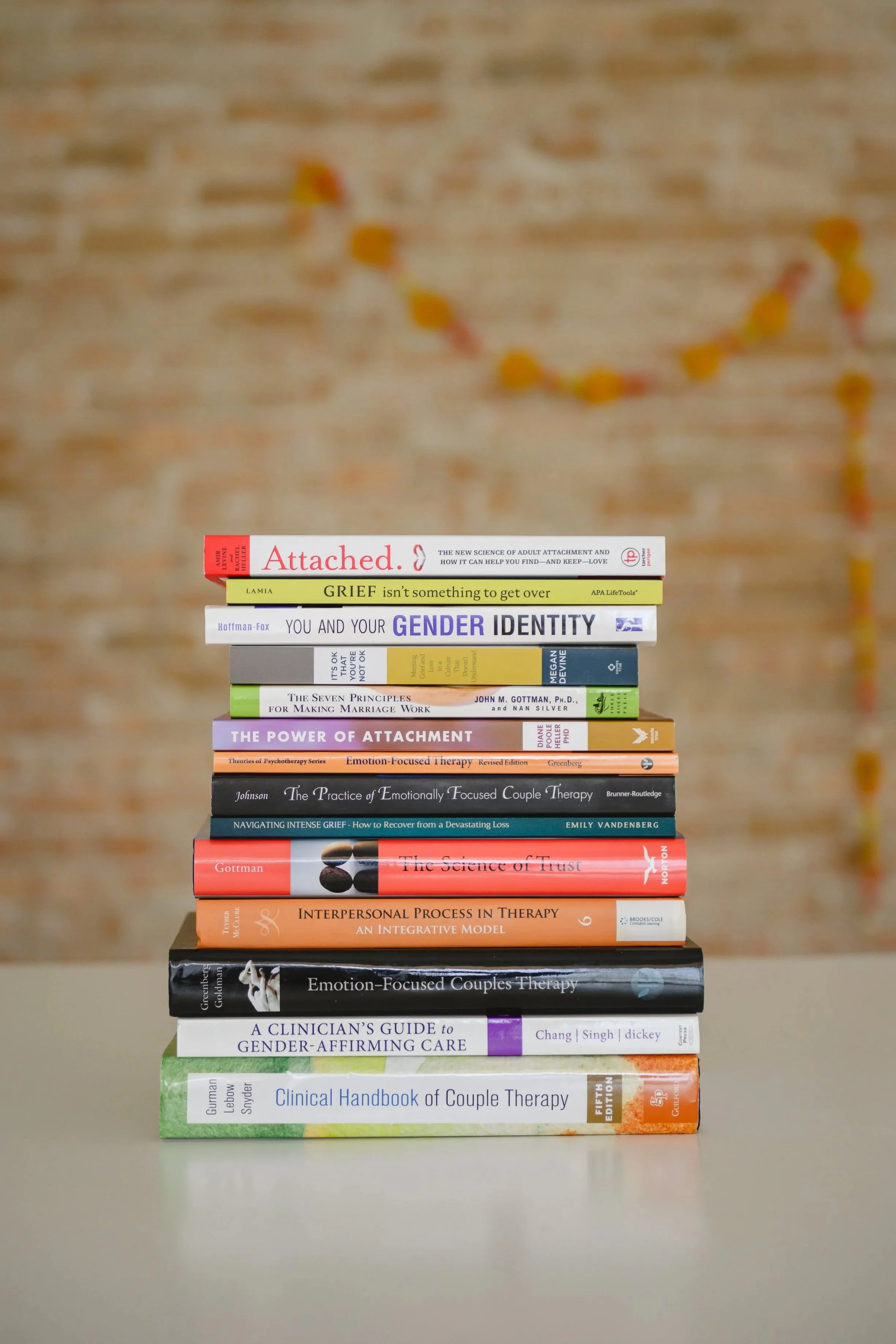 Stack of books on therapy and relationships placed on a table, with a blurred background of a brick wall with a string of orange and yellow beads hanging.