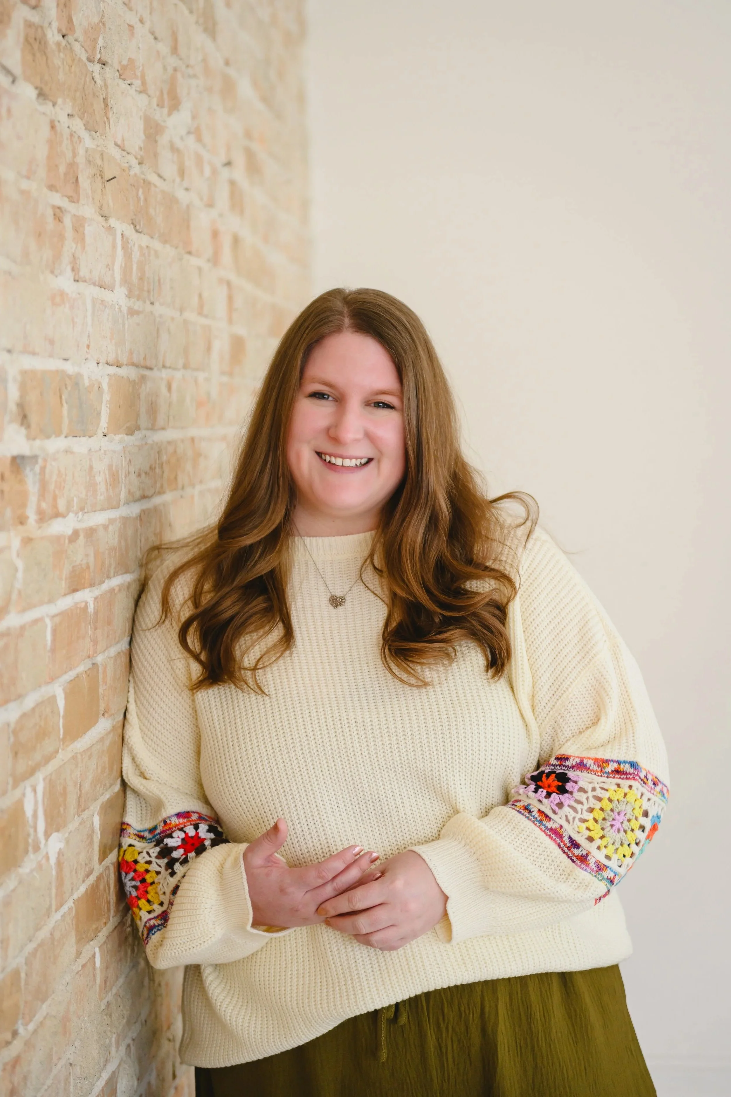 A woman with long, wavy red hair smiling while leaning against a brick wall, wearing a cream sweater with colorful embroidery on the sleeves and a necklace.