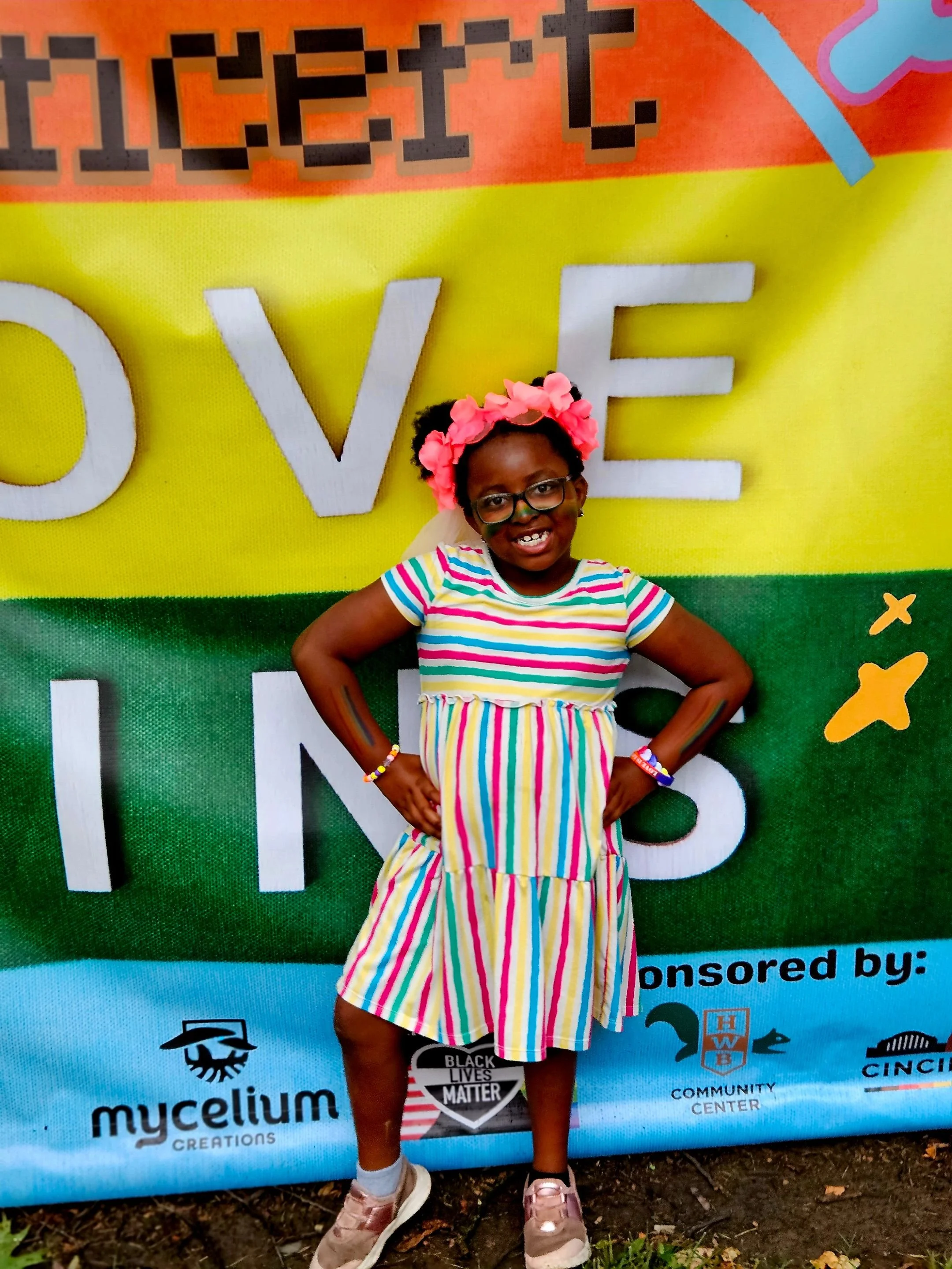 A young person of color stands in front of a rainbow sign that says "Love Wins." The bottom-right corner lists the names of our local partners who made the neighborhood event possible.