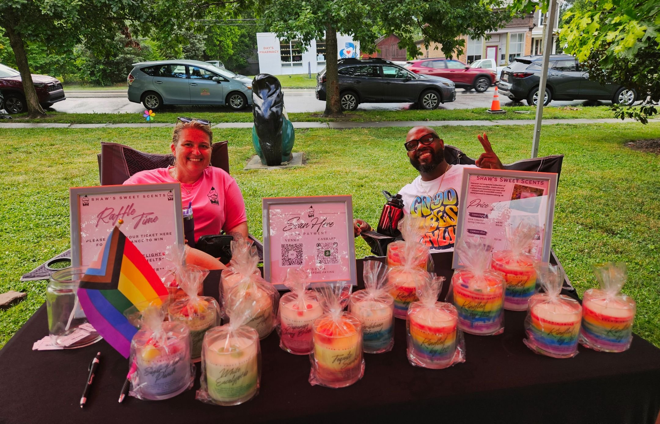 A Local Vendor from Cincinnati selling multicolored candles. There is a man and a woman sitting in the background. The man is giving the photographer a peace sign.