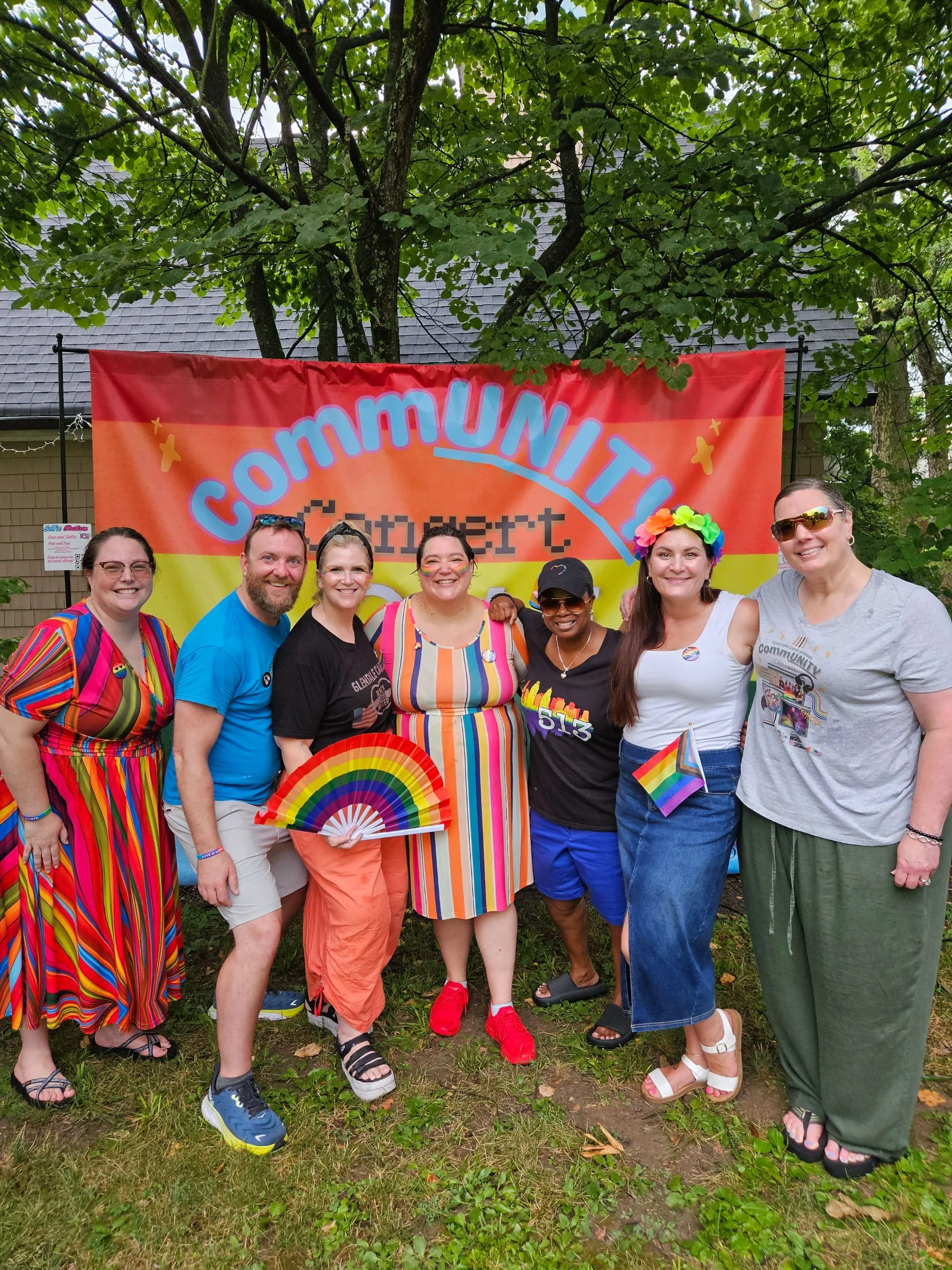 A group of 7 men and women of diverse backgrounds standing in front of a large rainbow colored sign that says "CommUNITY Concert, Love Wins." This group is the first organizers of the event.