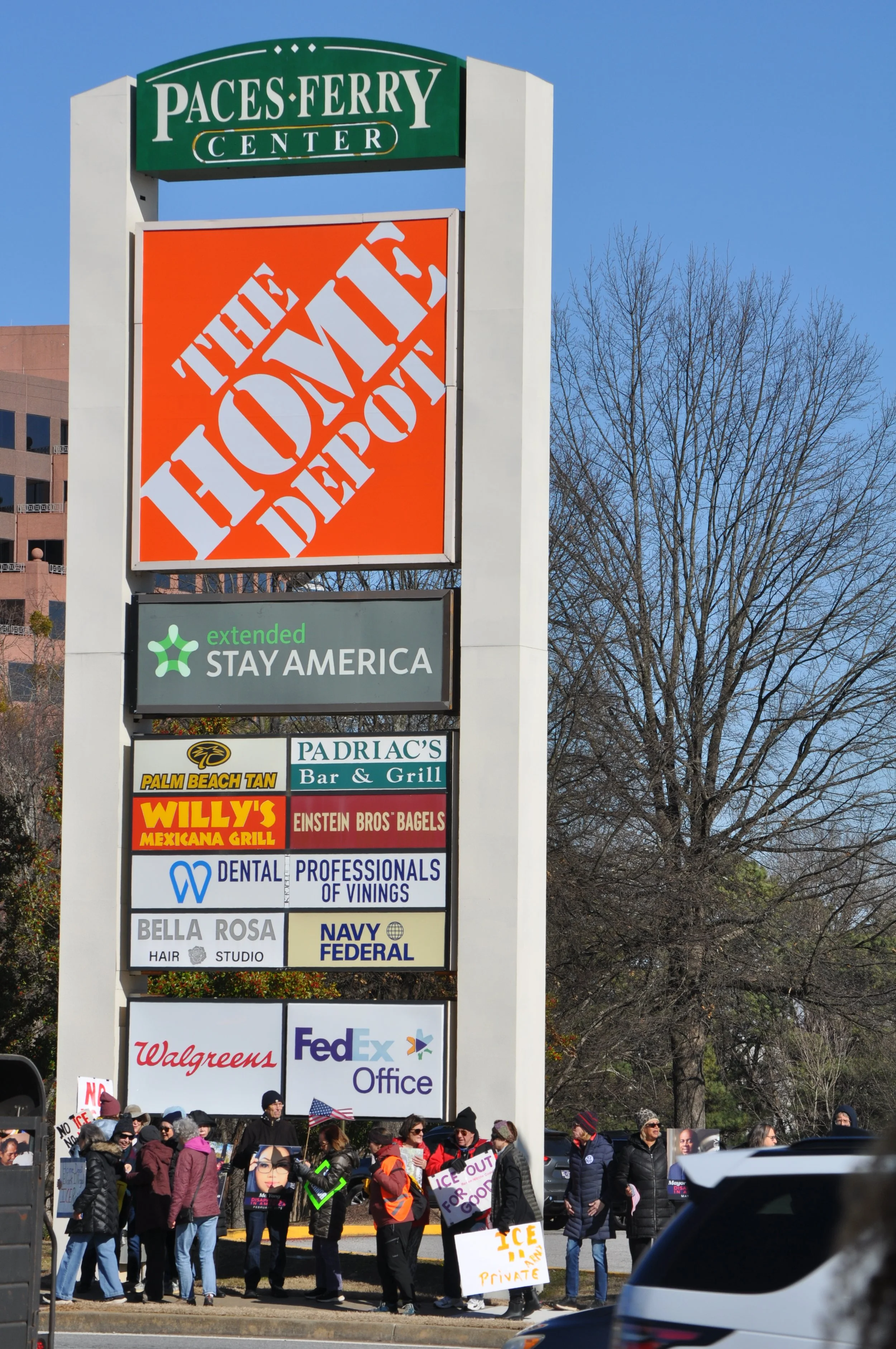 People protesting in front of a shopping center sign for Paces Ferry Center, The Home Depot, and various businesses.