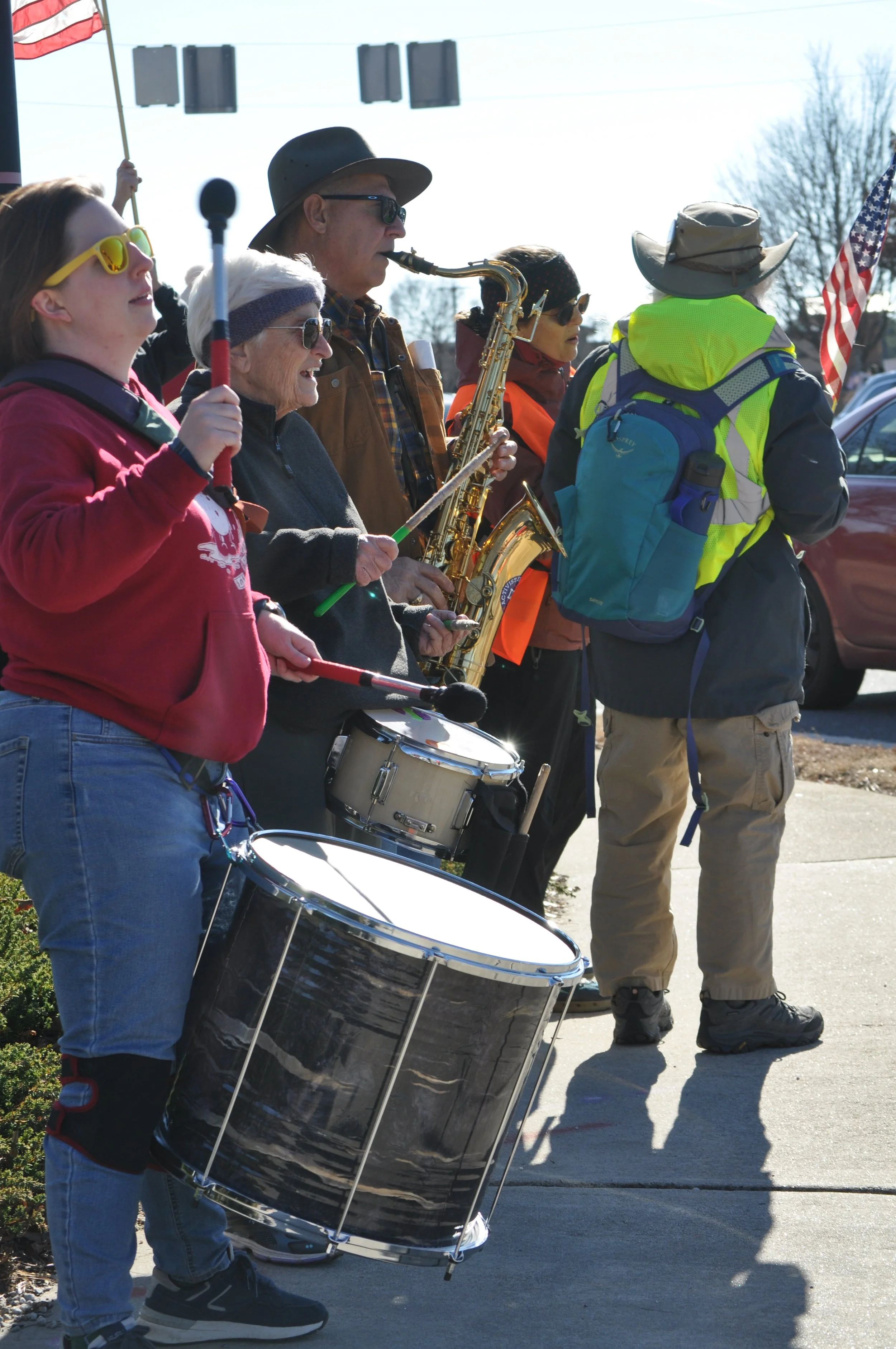 Group of people participating in a parade or outdoor event, playing musical instruments such as drums, saxophone, and other wind instruments, with some holding flags, standing on a sidewalk.