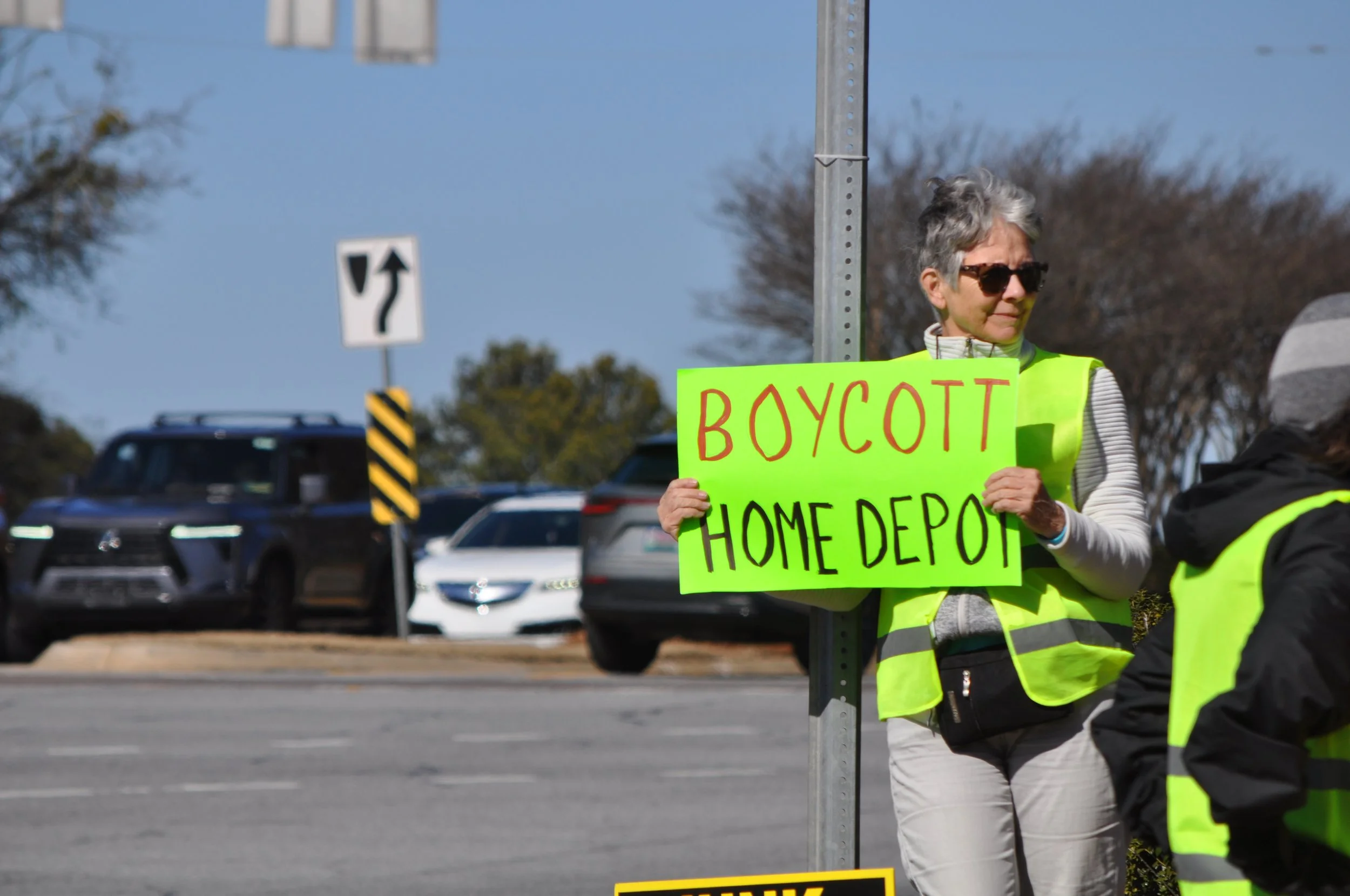 Woman holding a bright green protest sign that reads 'Boycott Home Depot' stands by a pole, with cars and a traffic sign in the background.
