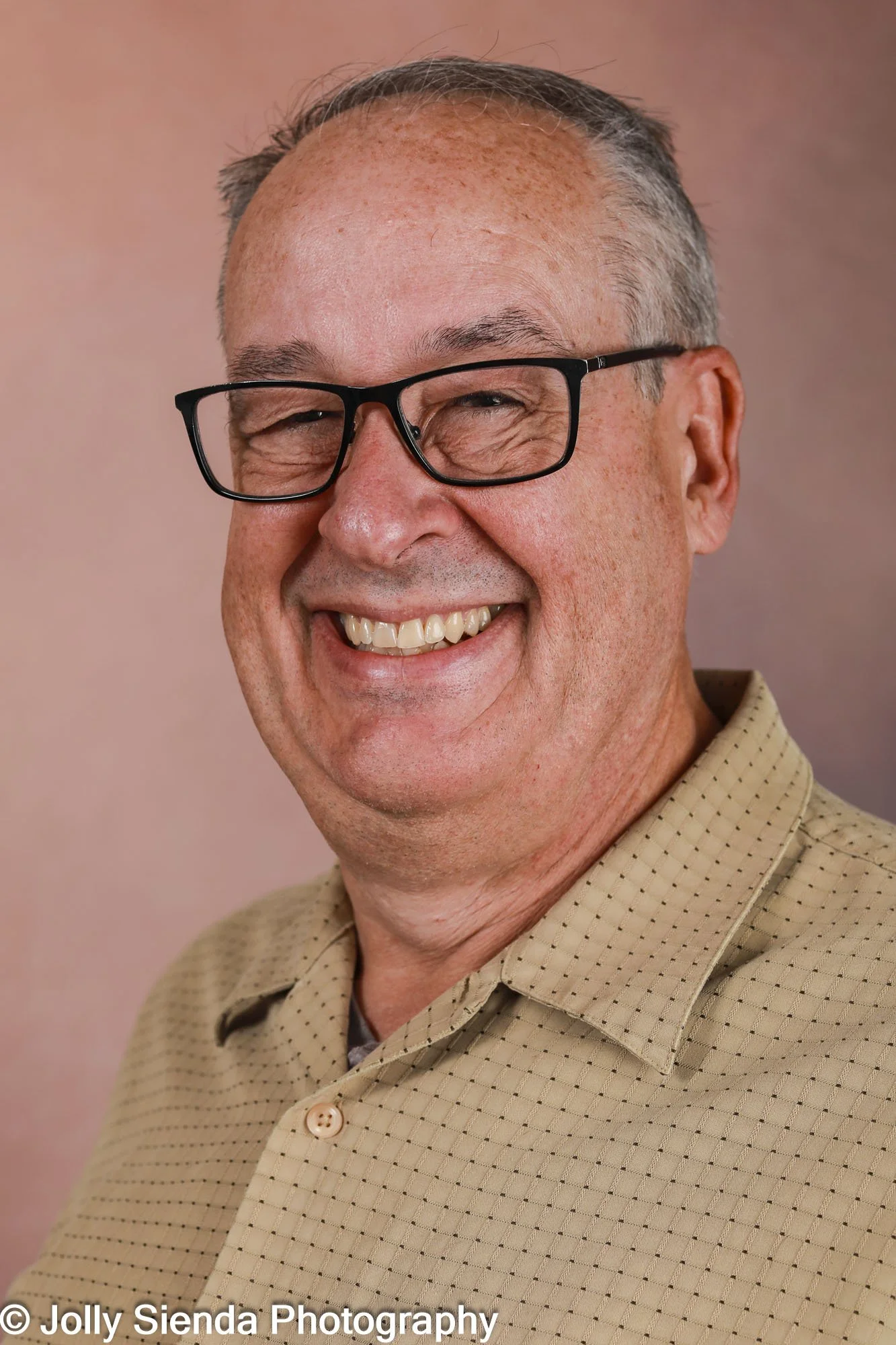 A middle-aged man with glasses and short gray hair smiling broadly, wearing a beige collared shirt against a soft pink background.