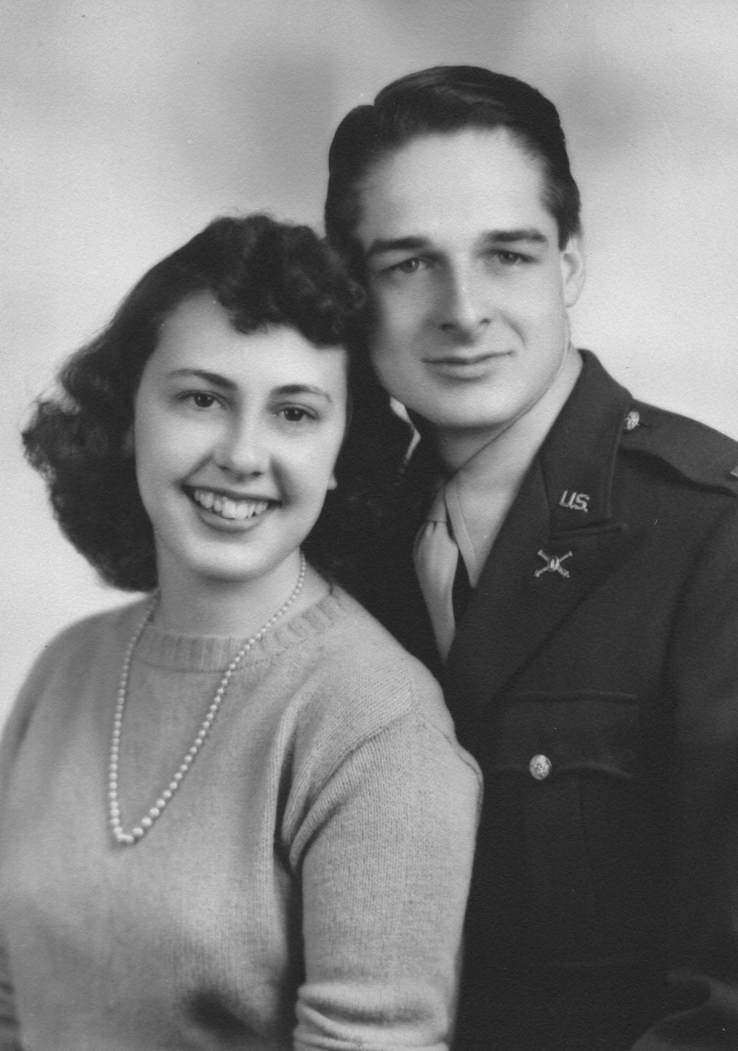 A black and white portrait of a smiling woman with dark, curly hair wearing a pearl necklace and a sweater, standing next to a young man in a military uniform.