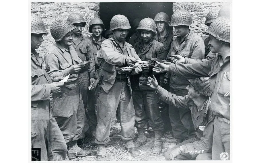 Black and white photo of a group of soldiers in military uniforms and helmets, with one soldier handing an item to another, surrounded by others, outdoors in a rocky area.
