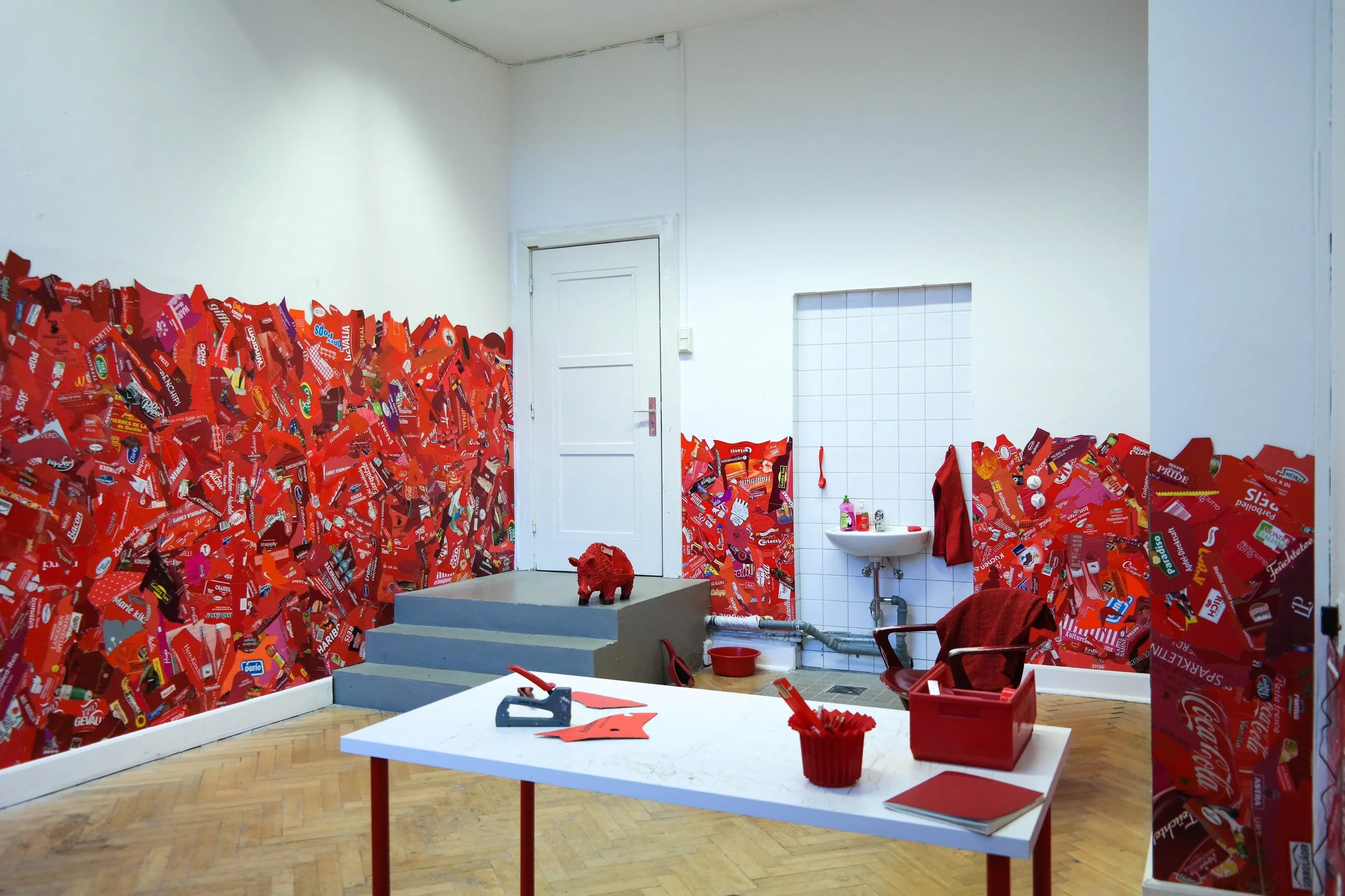Room decorated with red candy bar wrappers on the walls, a white table with craft supplies, a small stage with a red toy and steps, a sink area, and red chairs.
