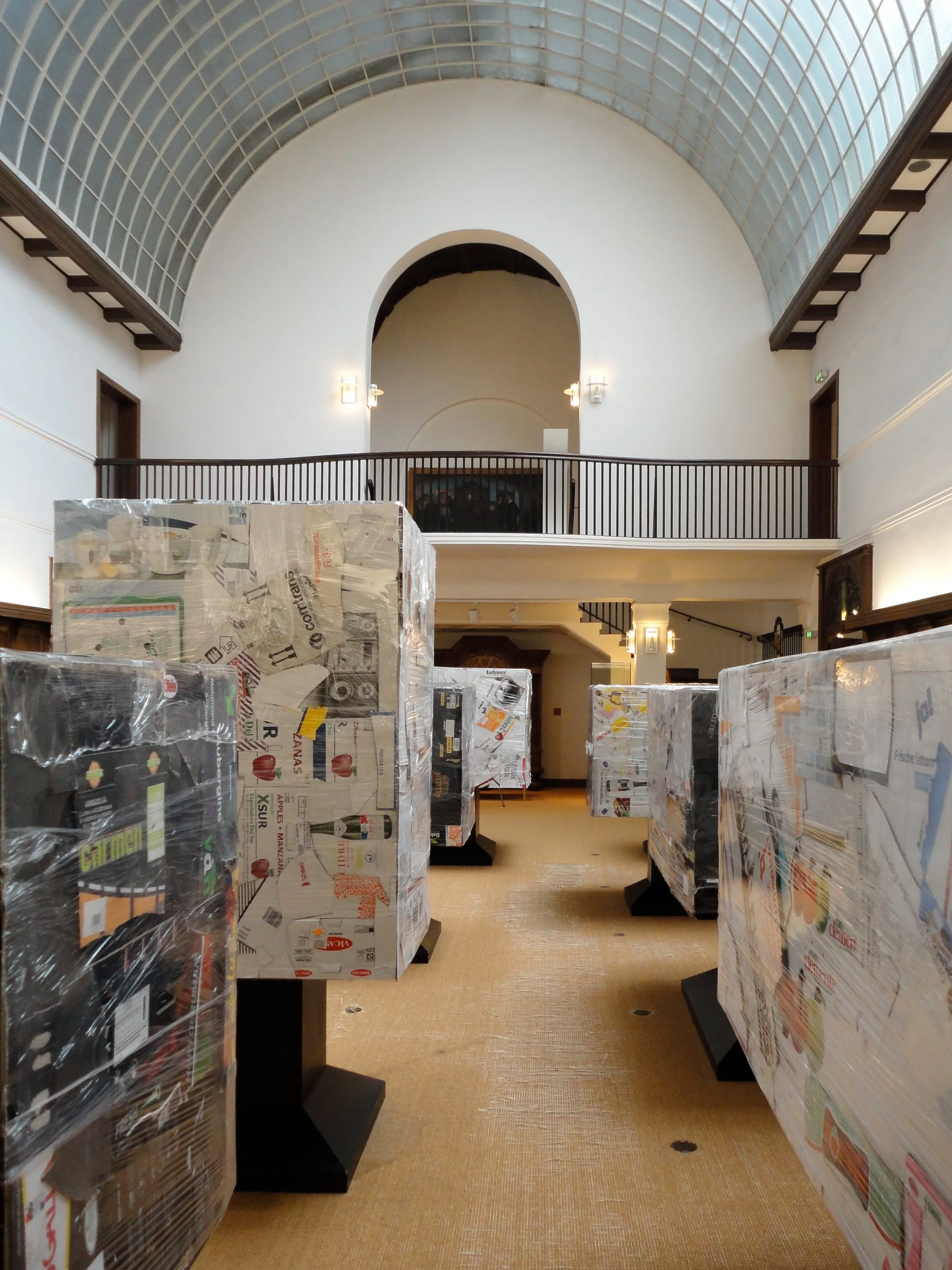 Indoor space with rows of display cases wrapped in plastic, a high arched ceiling with glass blocks, and a balcony with a black railing.
