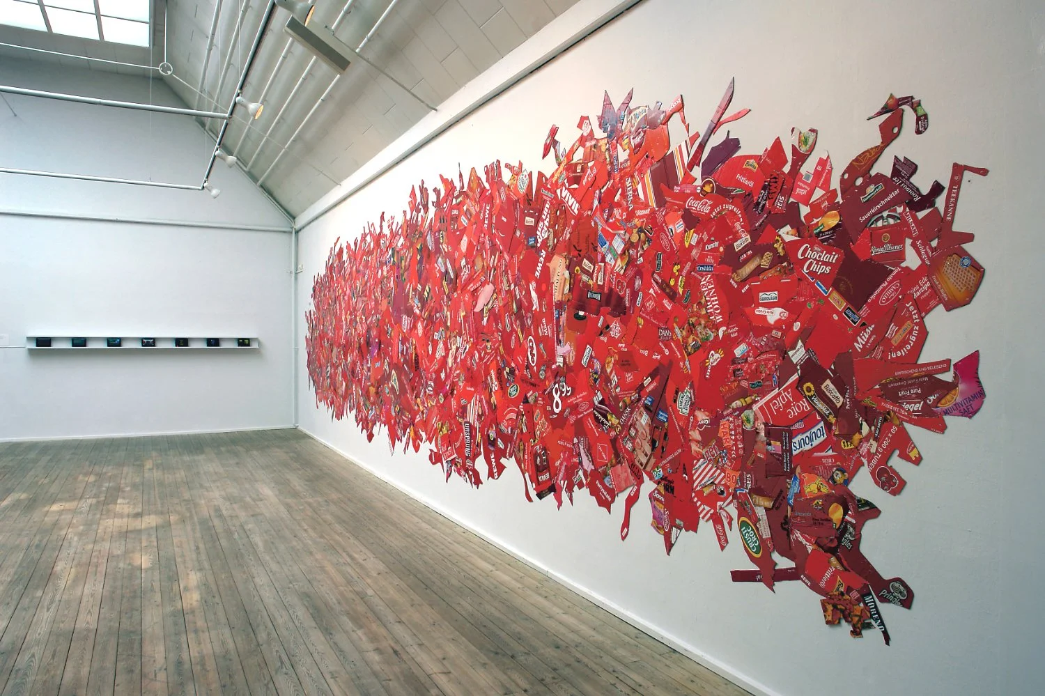 A large art installation on a white gallery wall made from cut-out candy box shapes, arranged in the form of a red heart.