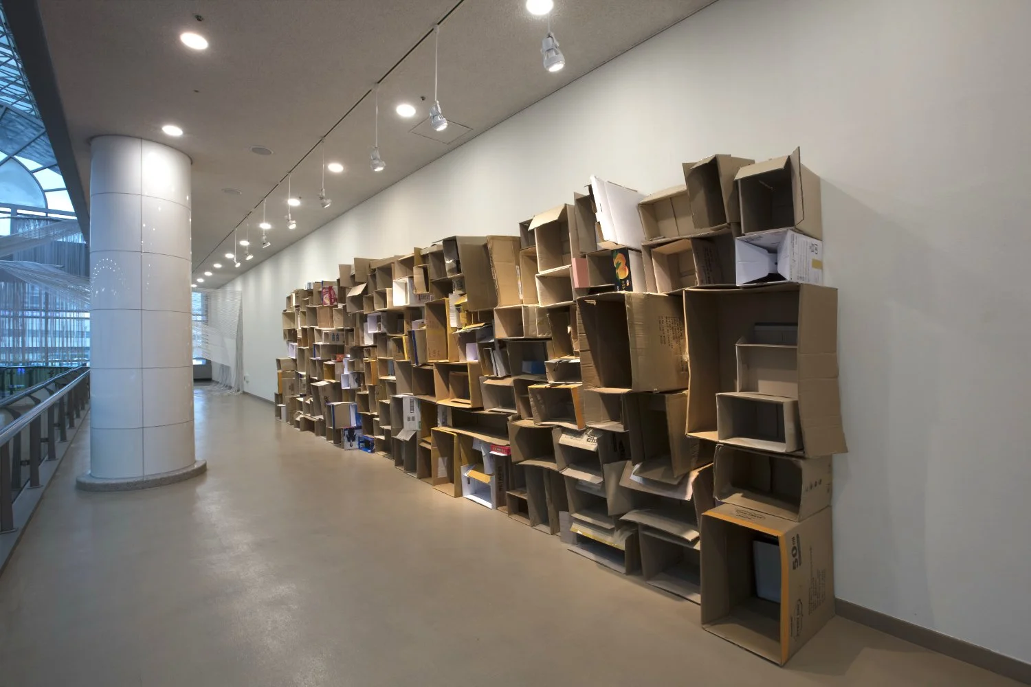 A hallway with a large wall of stacked cardboard boxes used as shelves, some containing books and other items. The hallway has a white ceiling with recessed lighting and a windowed wall on the left side.