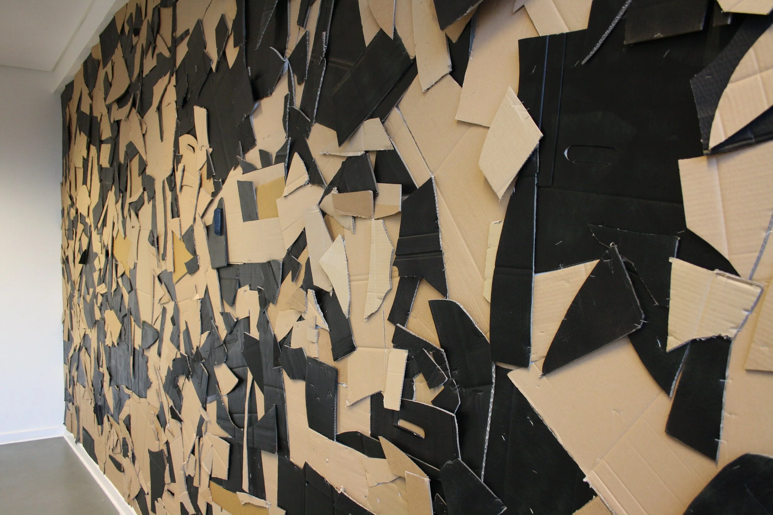 An abstract wall sculpture made from torn pieces of cardboard in black and beige colors arranged in a dynamic, layered pattern.