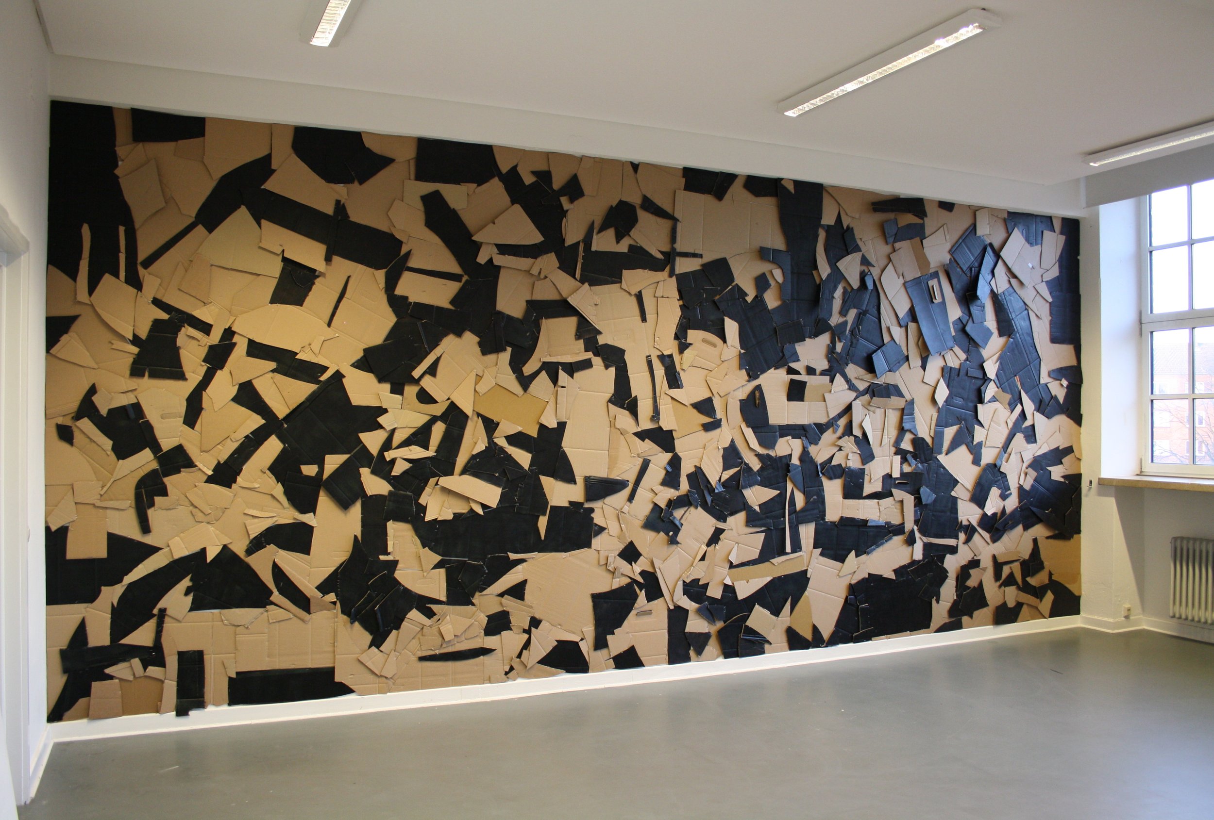 Wall covered with randomly arranged cardboard pieces in black and tan colors, in a well-lit room with a window on the right.