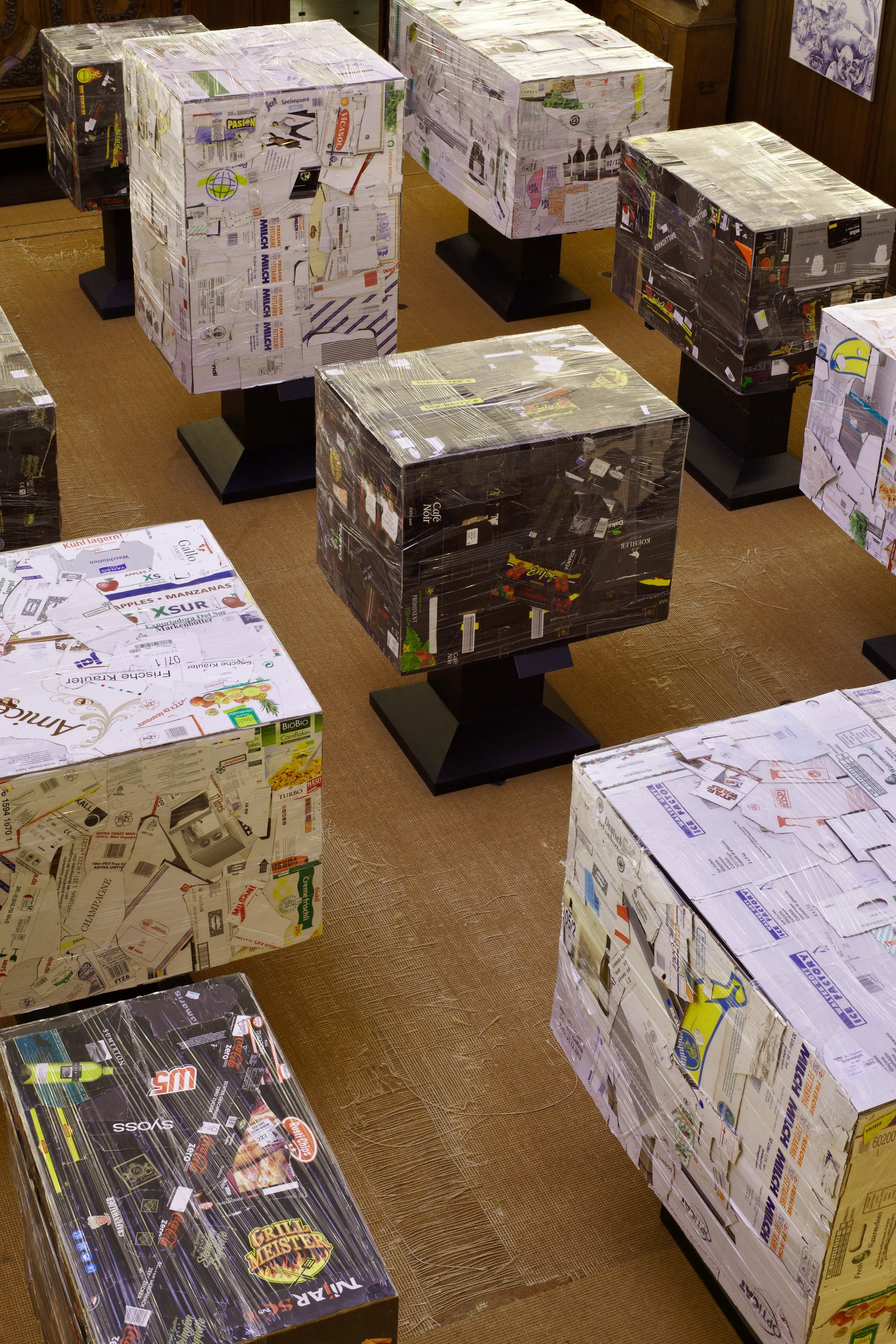 Multiple cardboard boxes covered with cardboard and plastic wrap, displayed on black stands in a room with brown carpet and wooden furniture.