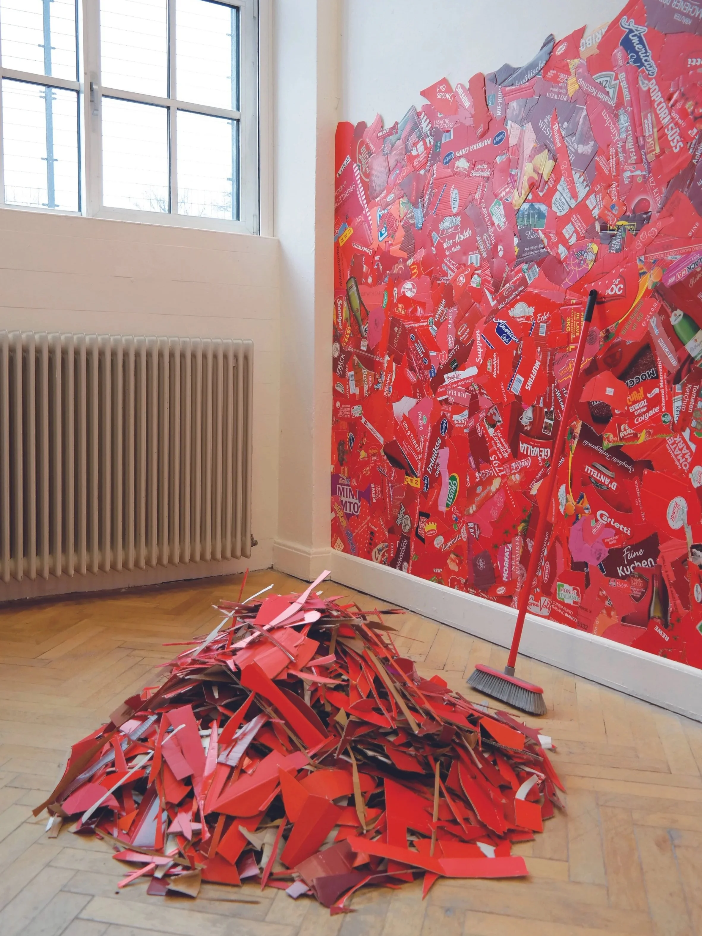 An art installation featuring a pile of torn red cardboard pieces on a wooden floor, a broom leaning against a wall, and a large wall covered in similarly torn red cardboard pieces in a room with a window and radiator.
