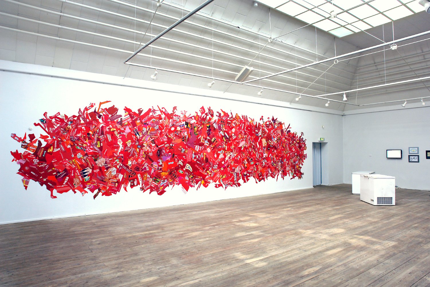 Large art installation on a white gallery wall made of numerous red, black, and yellow cutouts resembling skateboard decks with graphic designs. The gallery has a wooden floor, high ceilings with track lighting, and a few framed artworks on the adjacent wall.