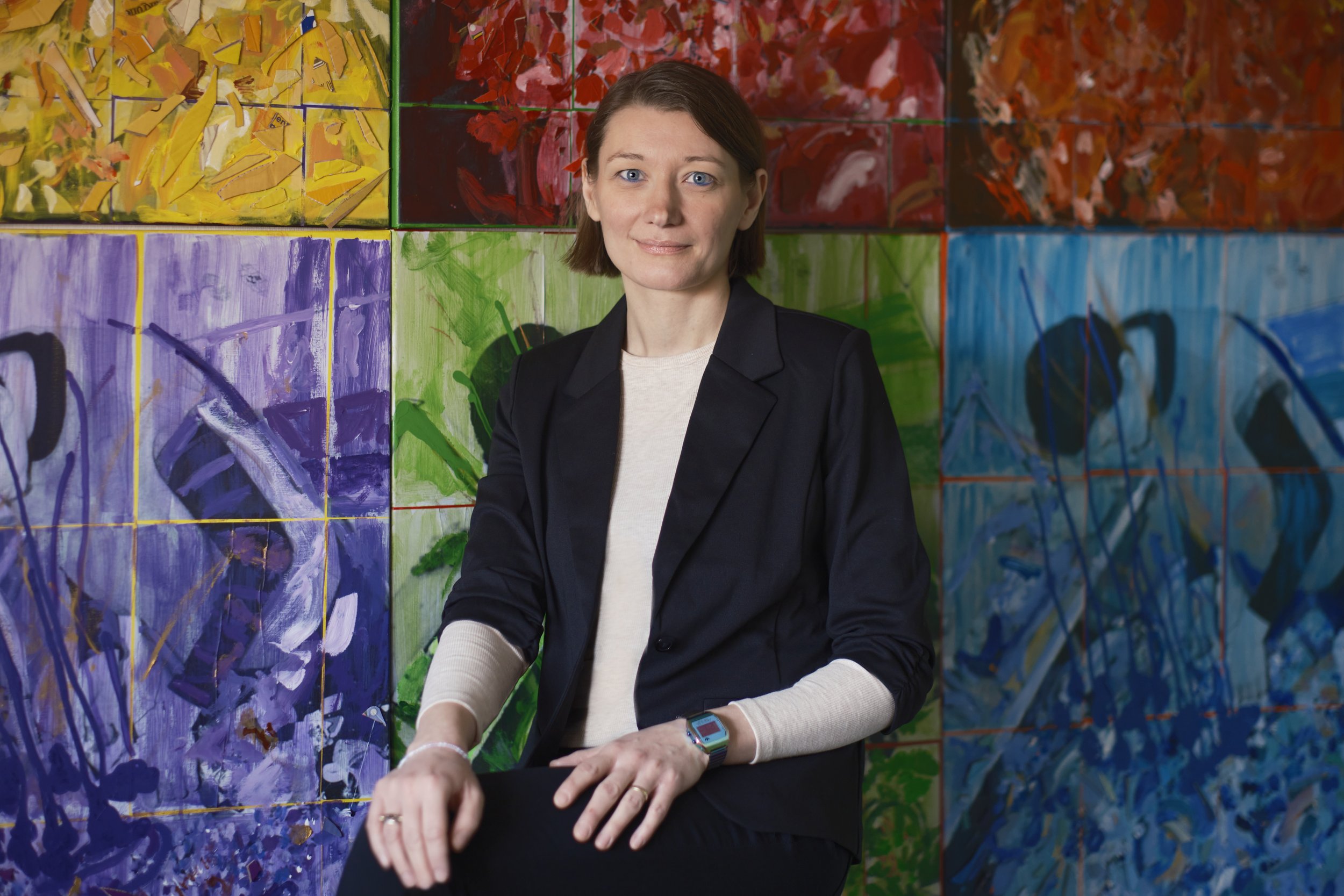 A woman with short brown hair and blue eyes, wearing a black blazer, sitting in front of a colorful abstract mural with various brushstrokes and colors. Photo by © Davy Denke