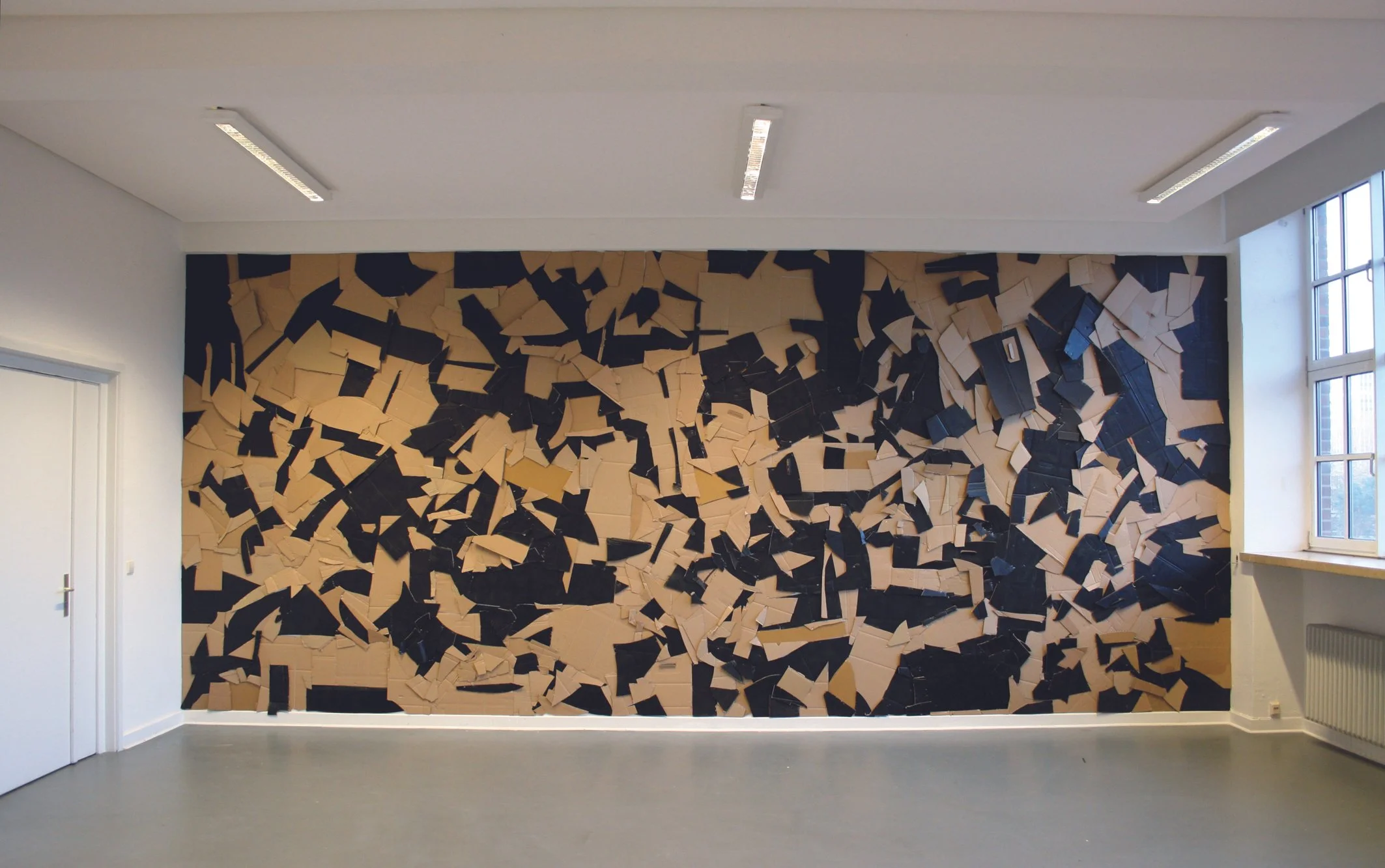 An abstract wall art installation with irregularly shaped pieces of cardboard painted in beige, black, and dark blue, arranged in a chaotic pattern in a room with white walls and large windows.