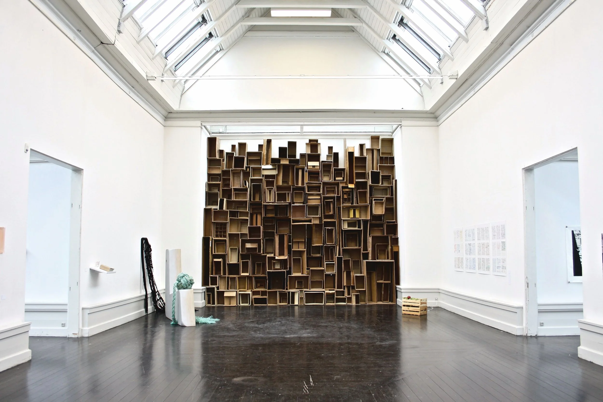 Interior view of an art gallery with a large cardboard bookshelf installation on the back wall, dark hardwood floor, white walls, and skylights in the ceiling.