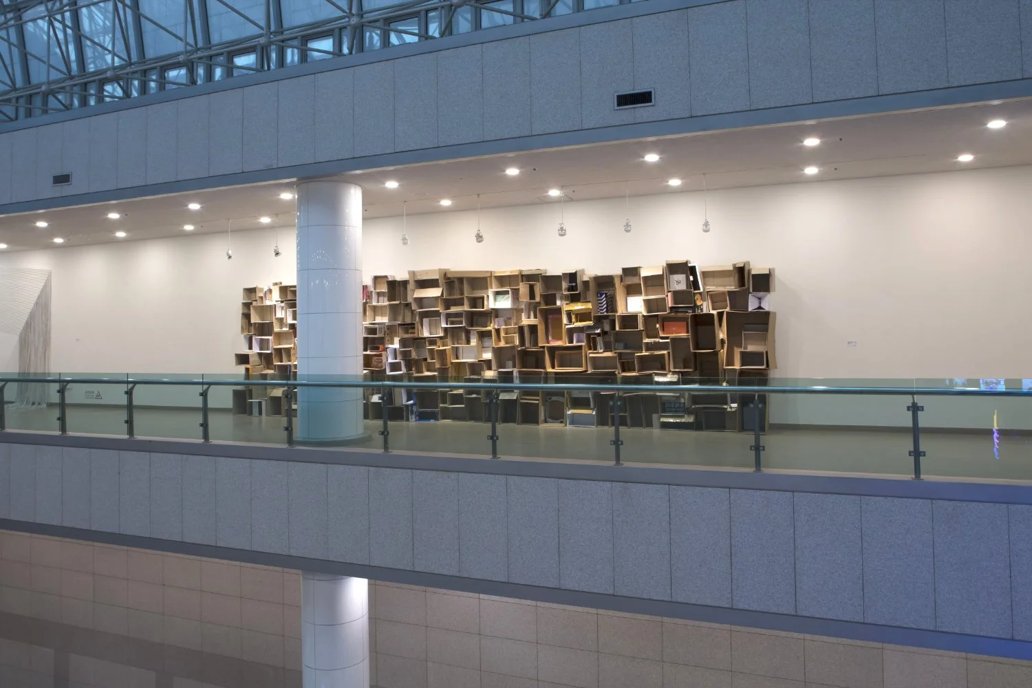 Interior of a modern building with a large art installation made of wooden boxes attached to the wall, glass railing, white walls, and ceiling lights.