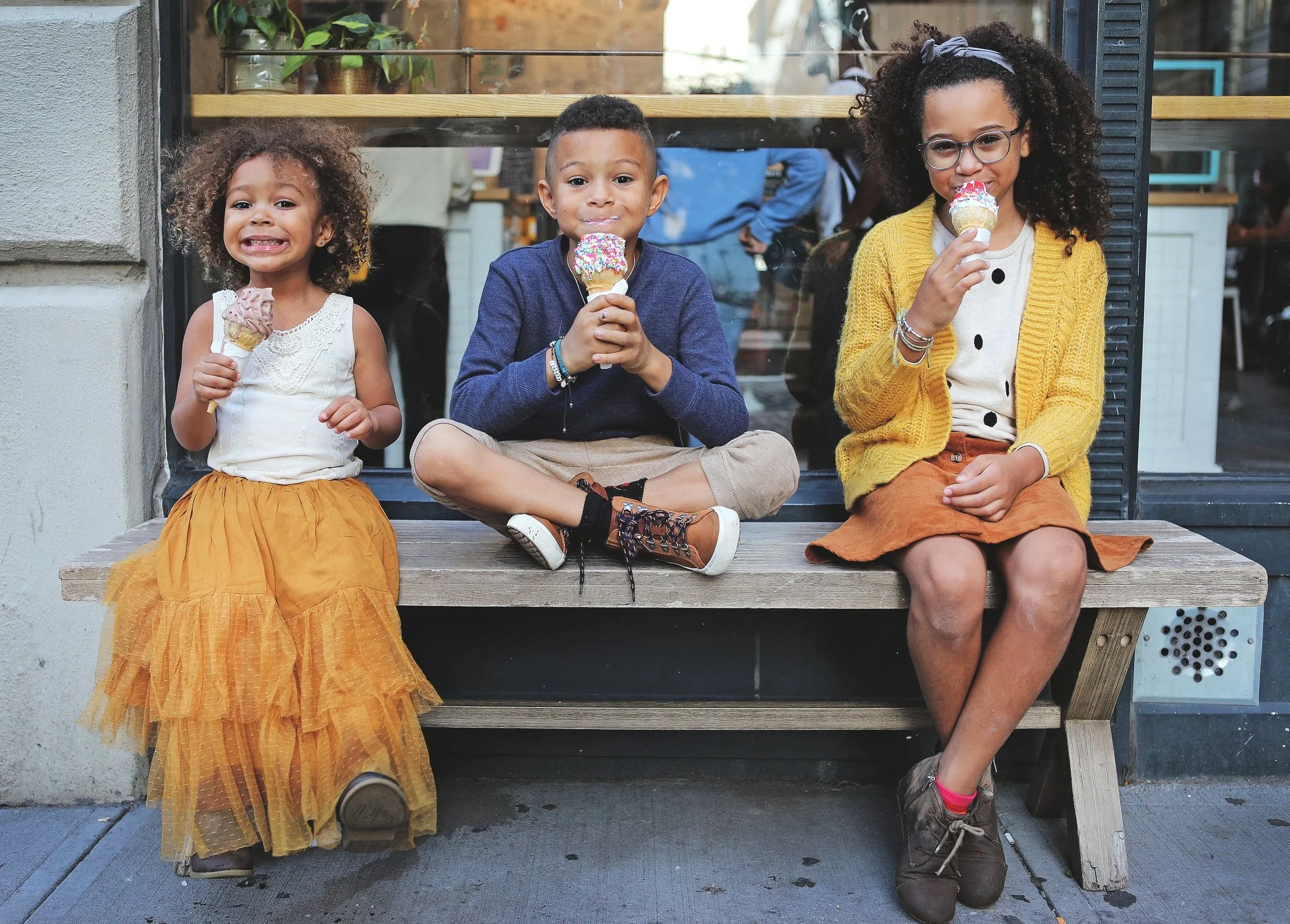 Three children sitting on a wooden bench outside, each holding an ice cream cone with colorful sprinkles, smiling and enjoying their treat.