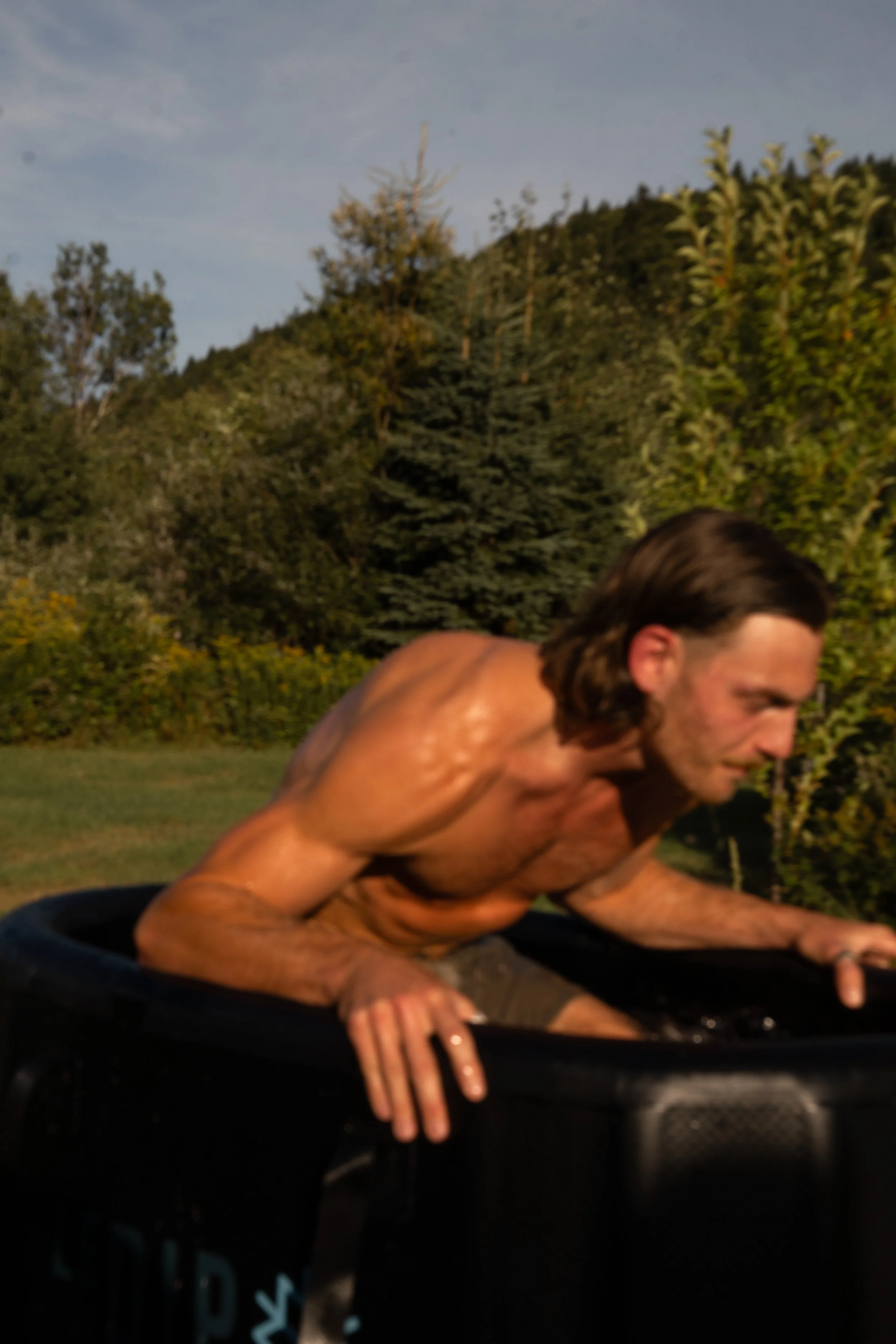 Un homme nu, avec de longs cheveux bruns, est à l'extérieur, probablement en train de faire du yoga ou une autre activité physique, avec un paysage verdoyant et un ciel clair en arrière-plan.