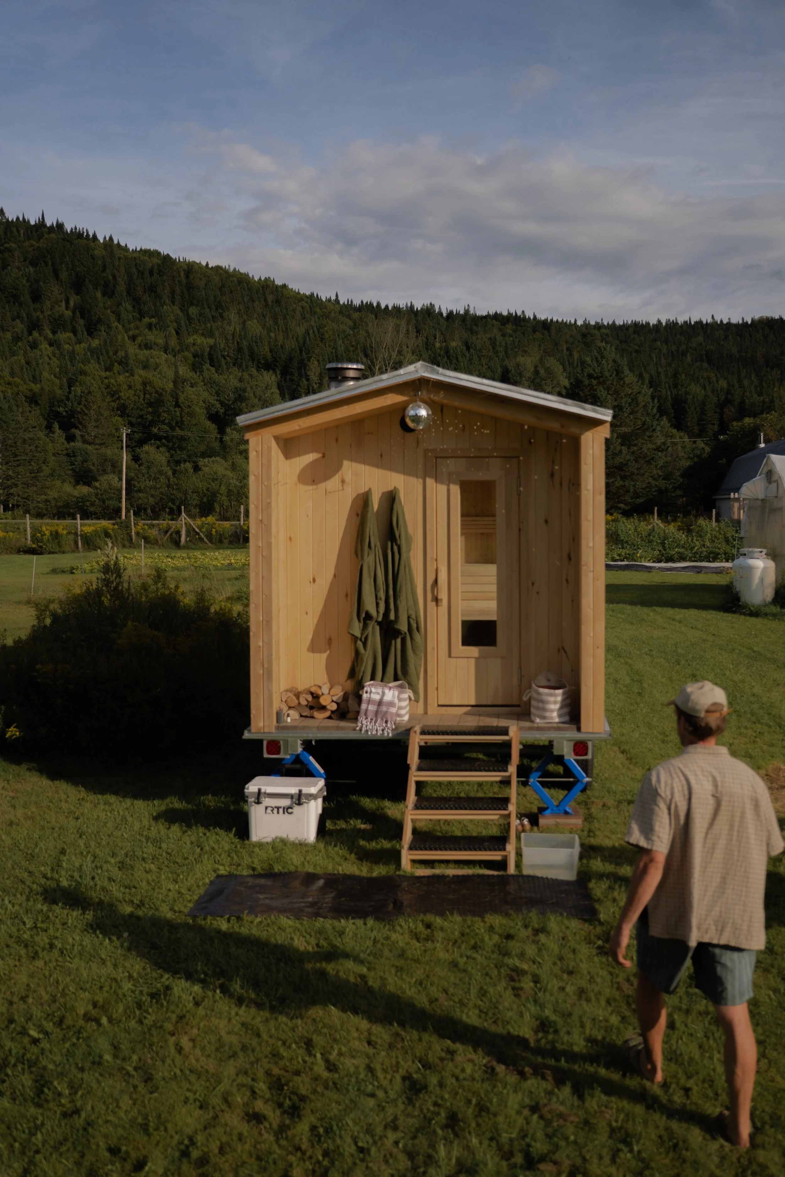 Petit sauna en bois sur une remorque dans un jardin avec un homme marchant à proximité, entouré d'une nature verdoyante et d'une montagne au fond, sous un ciel partiellement nuageux.