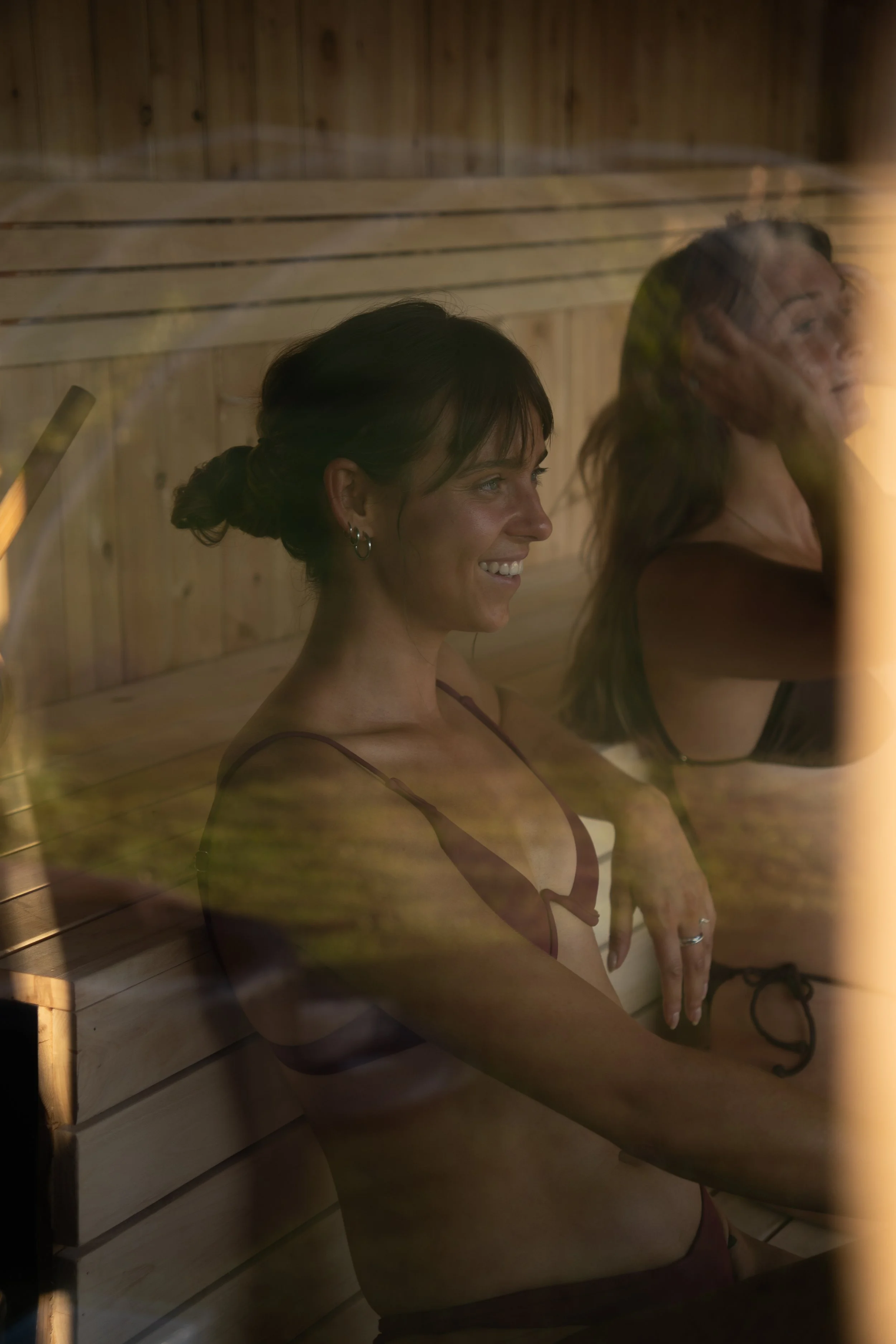Deux femmes assises dans un sauna en bois, l'une souriante, portant une robe noire ou un maillot de bain rouge, l'autre de profil avec des lunettes. La lumière chaude crée une ambiance relaxante.