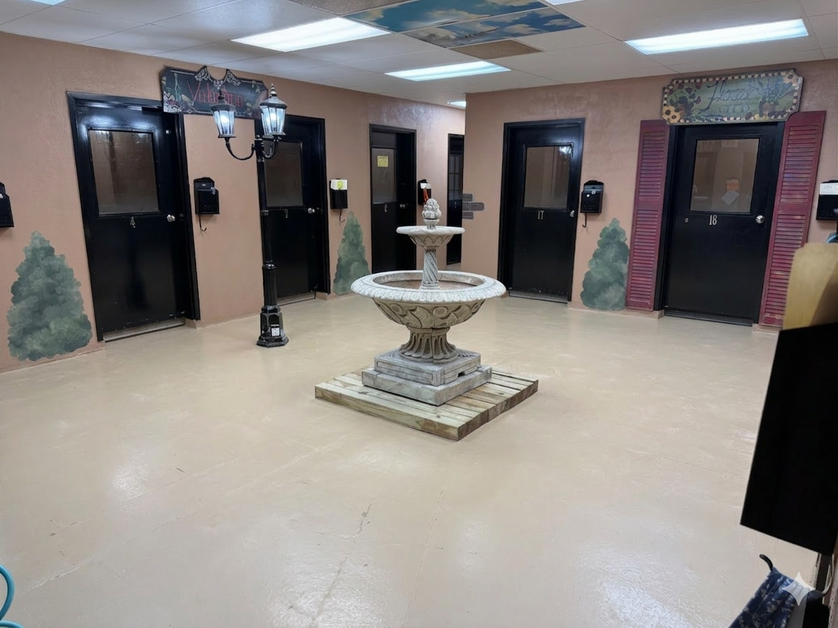 Interior of a building lobby with beige walls, black doors numbered 17 and 18, and murals of trees painted on the walls. A stone fountain is in the center of the room, and a small decorative lamp post stands near the wall.