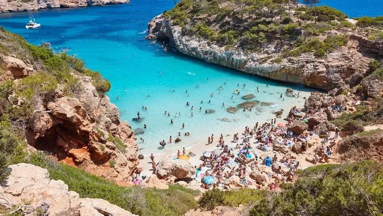 Crowded beach with turquoise water, surrounded by rocky cliffs and green vegetation, people swimming and sunbathing.
