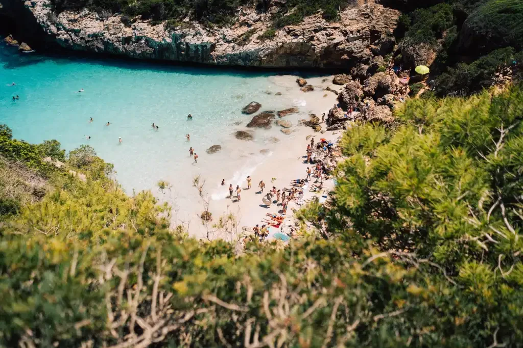 A beach with clear turquoise water surrounded by rocky cliffs and green foliage, filled with people swimming and relaxing on the sand.