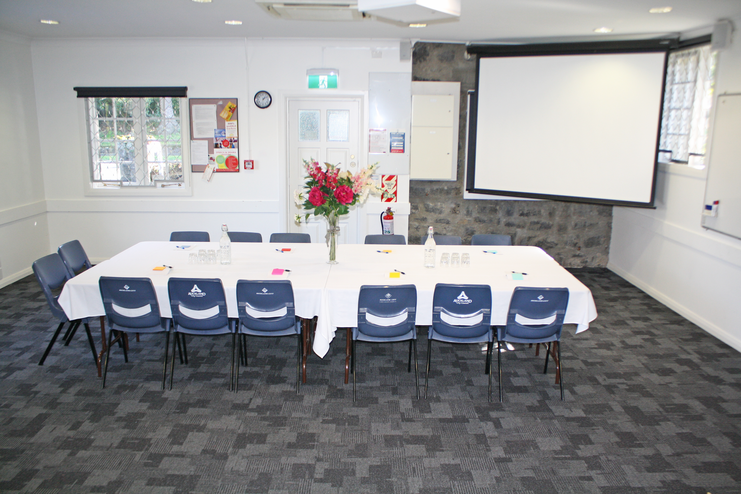 Meeting room with a rectangular table covered with a white cloth, set with bottles of water, glasses, surrounded by blue chairs with Auckland City logos, a large floral centerpiece, a projection screen, and window with blinds.