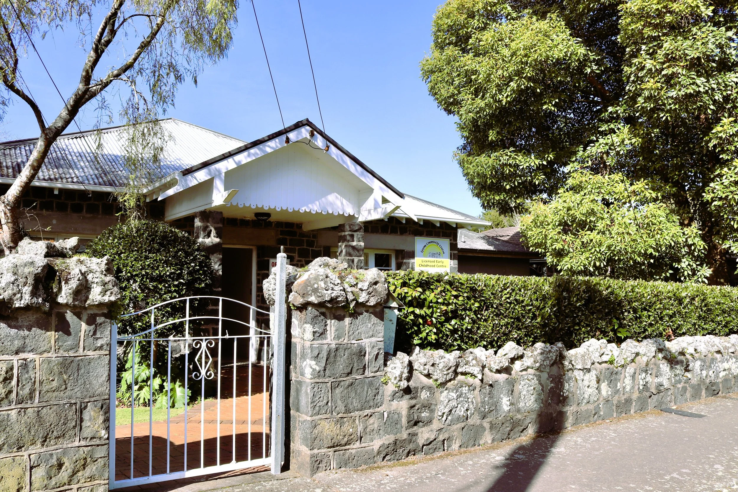 Entrance to Epsom Community center with a stone wall, white gate, and hedges, surrounded by trees under a blue sky.