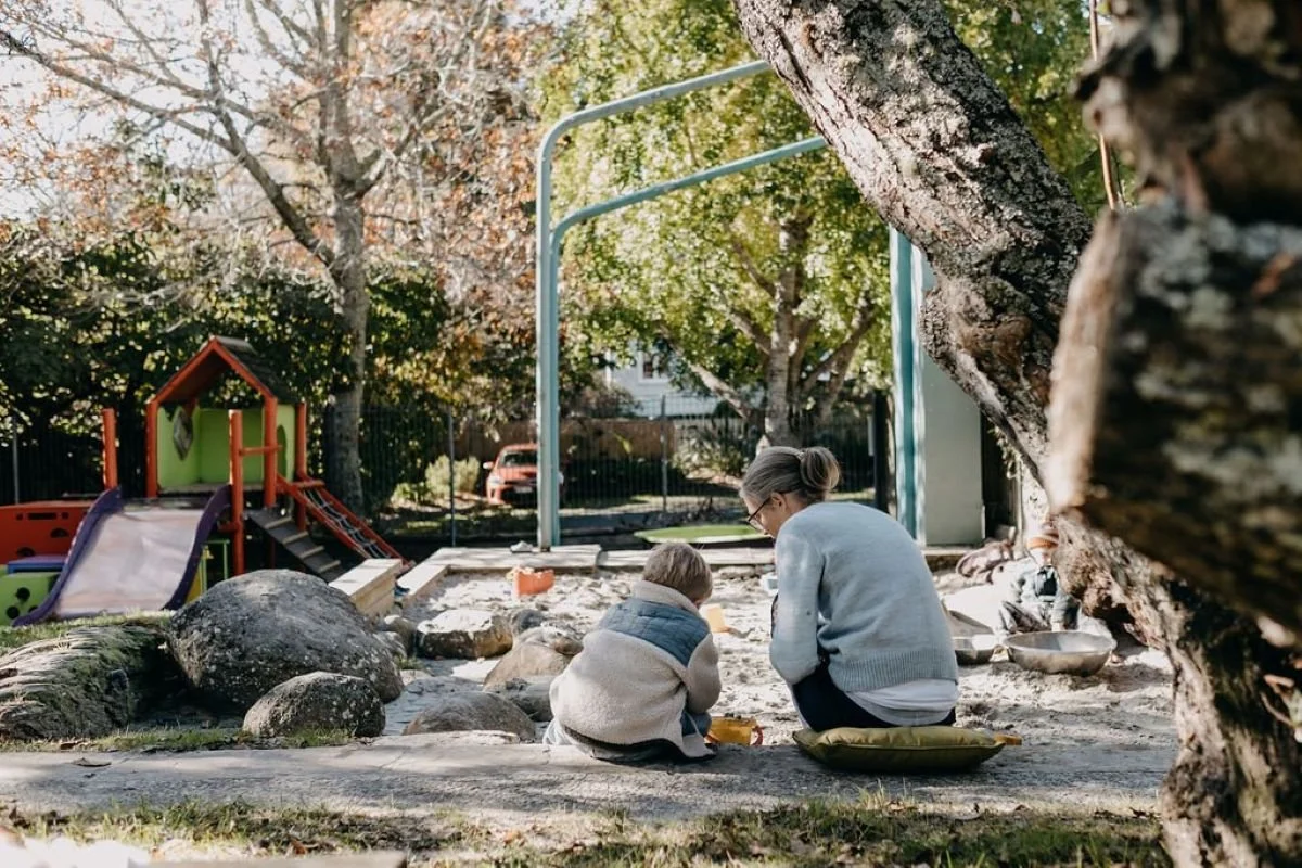 Teacher sits with child in the sandpit, child is digging in the sand with a bucket. Green trees and playground in the background.