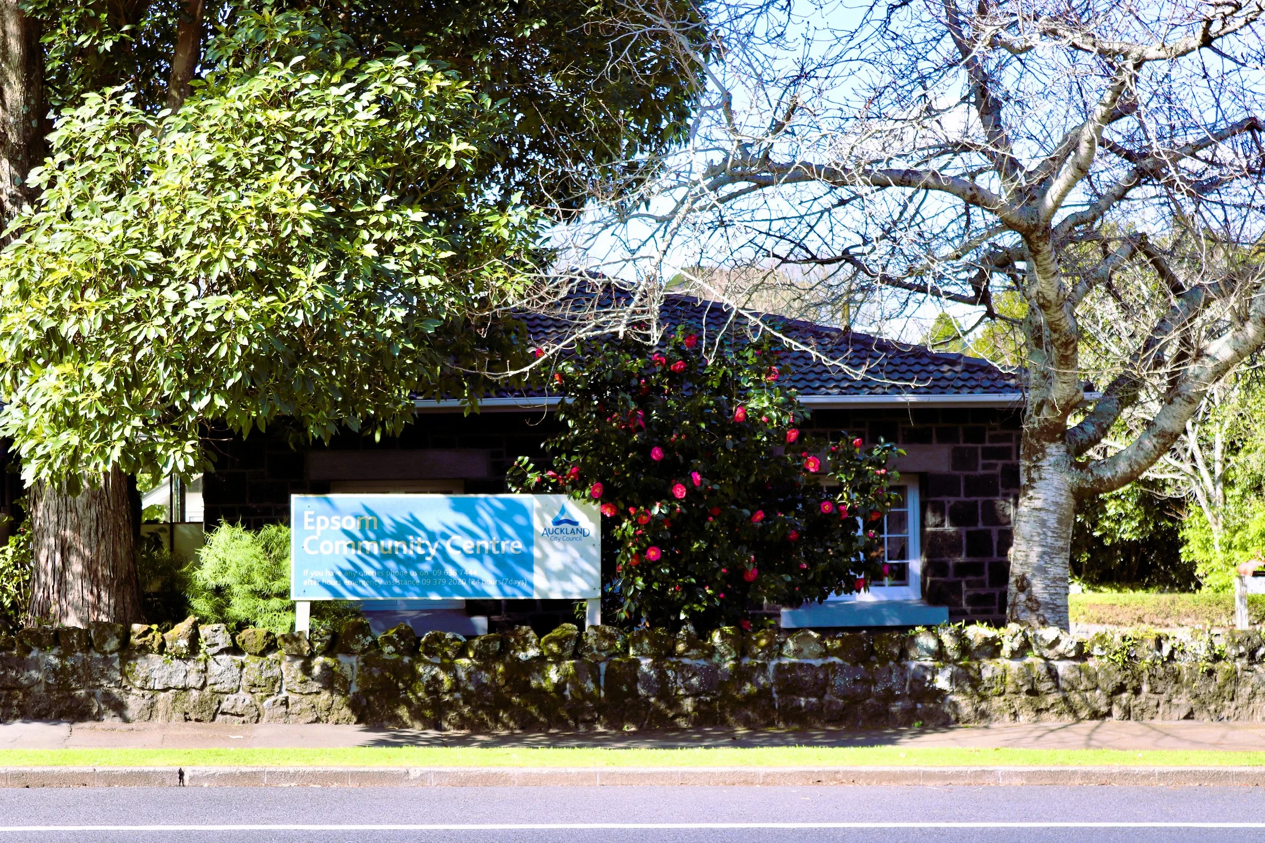An historical stone building with a sign that reads 'Epsom Community Centre,' surrounded by trees and bushes, with a stone wall in front and a roadway at the bottom.
