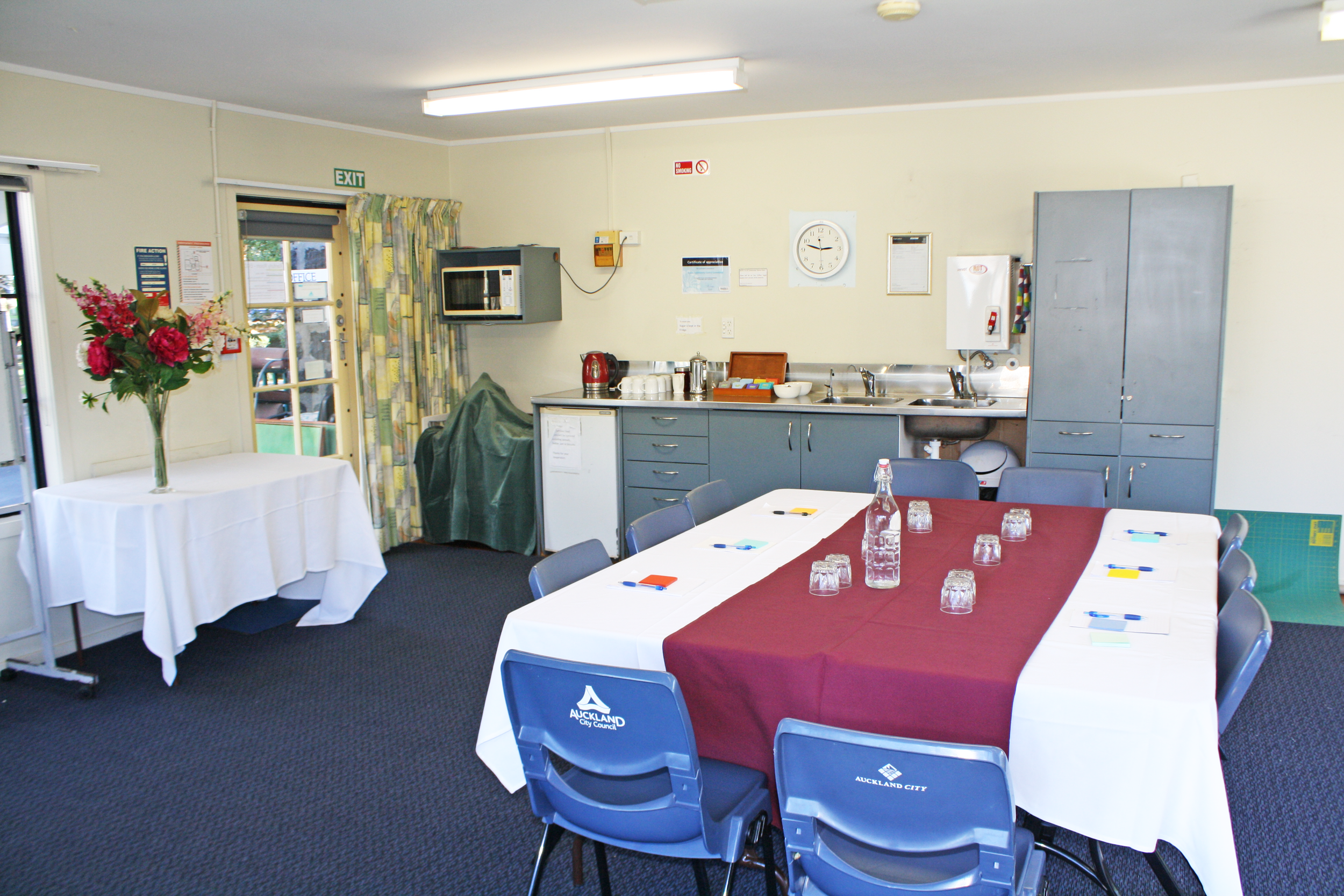 Meeting room with a rectangular table covered with a white tablecloth and a maroon tablecloth, surrounded by blue chairs with 'Auckland City' logo, set with water bottles, glasses, and notepads. A table with a flower vase is on the left. Kitchen area with cabinets, a sink, microwave, and coffee equipment is at the back. Exit door with a glass window and curtains is on the left. Fluorescent lights overhead and a wall clock are also visible.