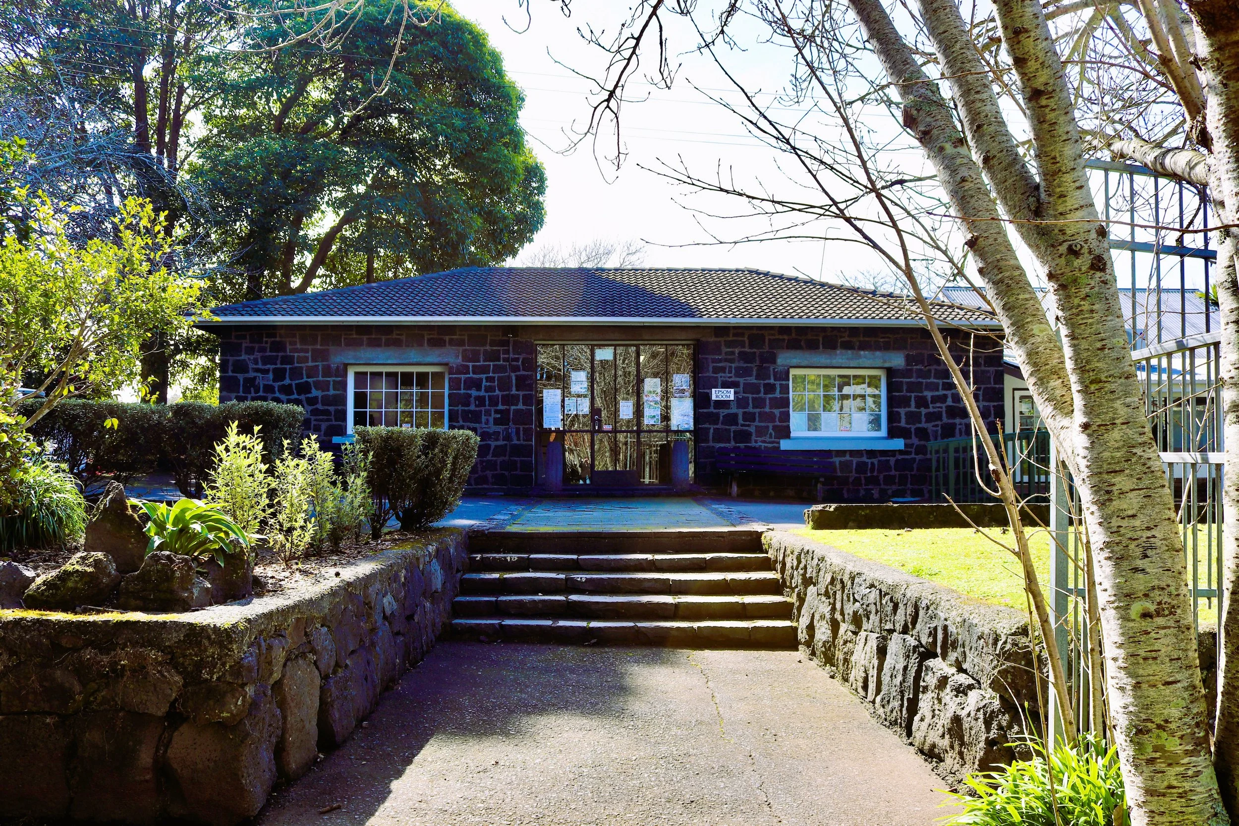 A stone building with a dark stone exterior, surrounded by trees and shrubs, with steps leading up to a glass door entrance.