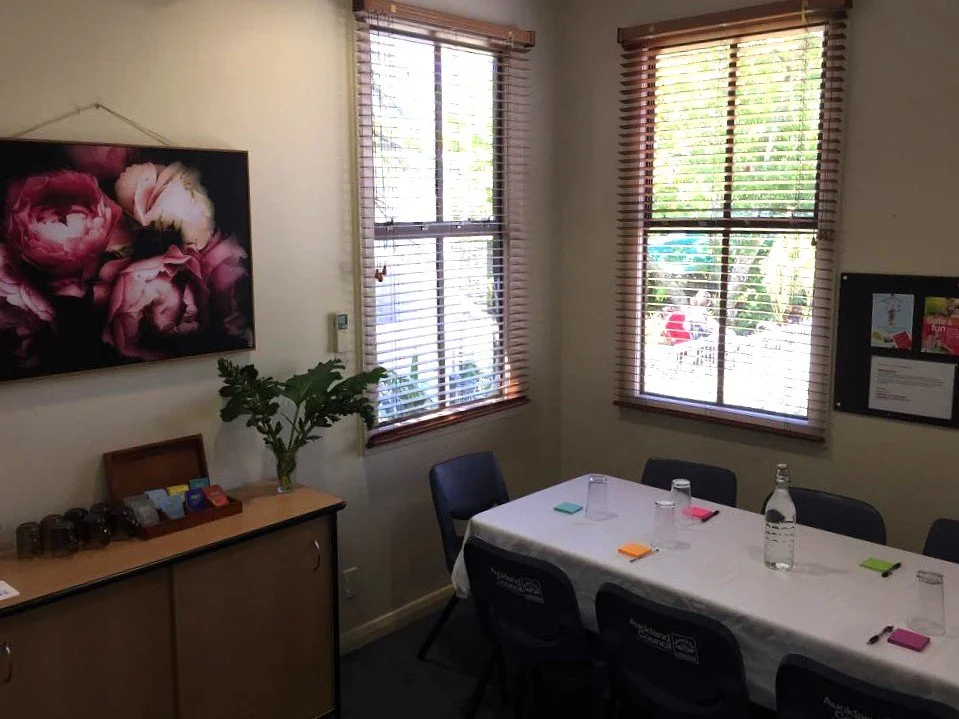 A meeting room with a rectangular table covered with a white tablecloth, six black chairs, three water bottles The room has two windows with wooden blinds, a large floral painting on the wall and a black bulletin board on the wall.
