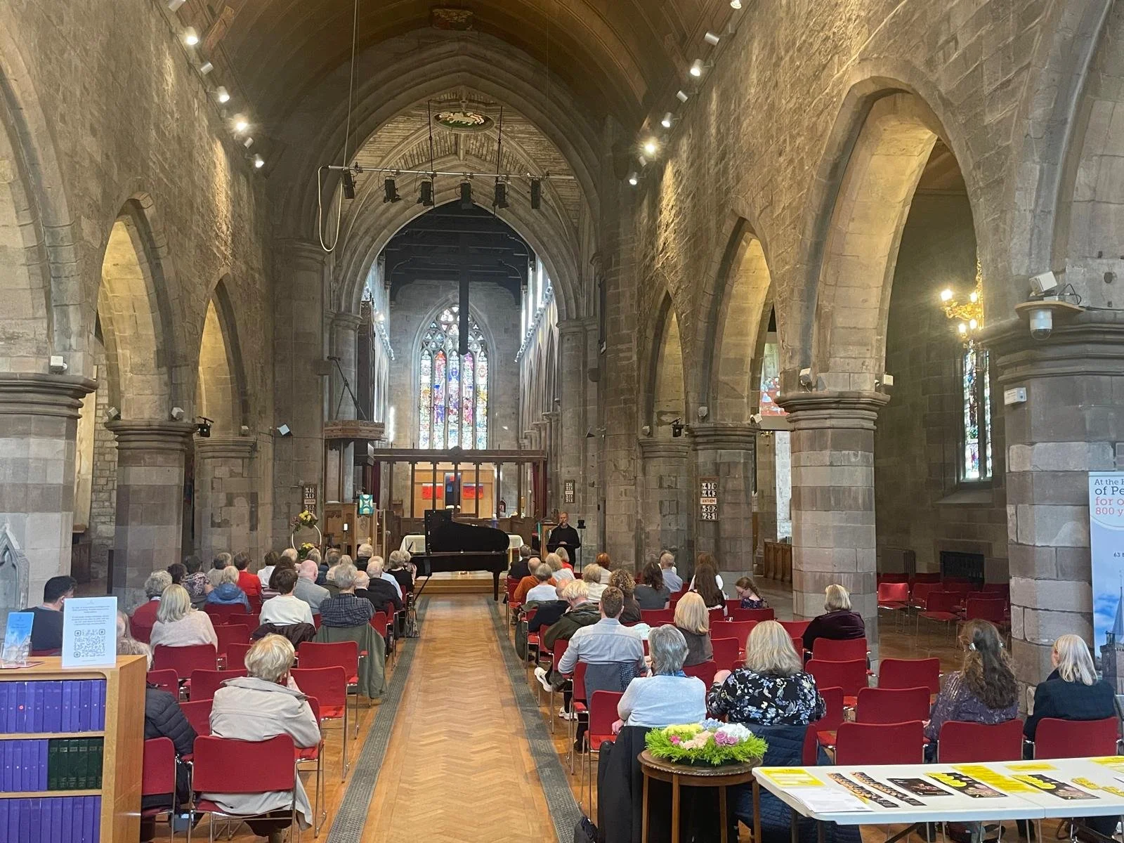People attending a musical performance inside a Gothic-style stone church with stained glass windows, a black grand piano, and an audience seated in red chairs.