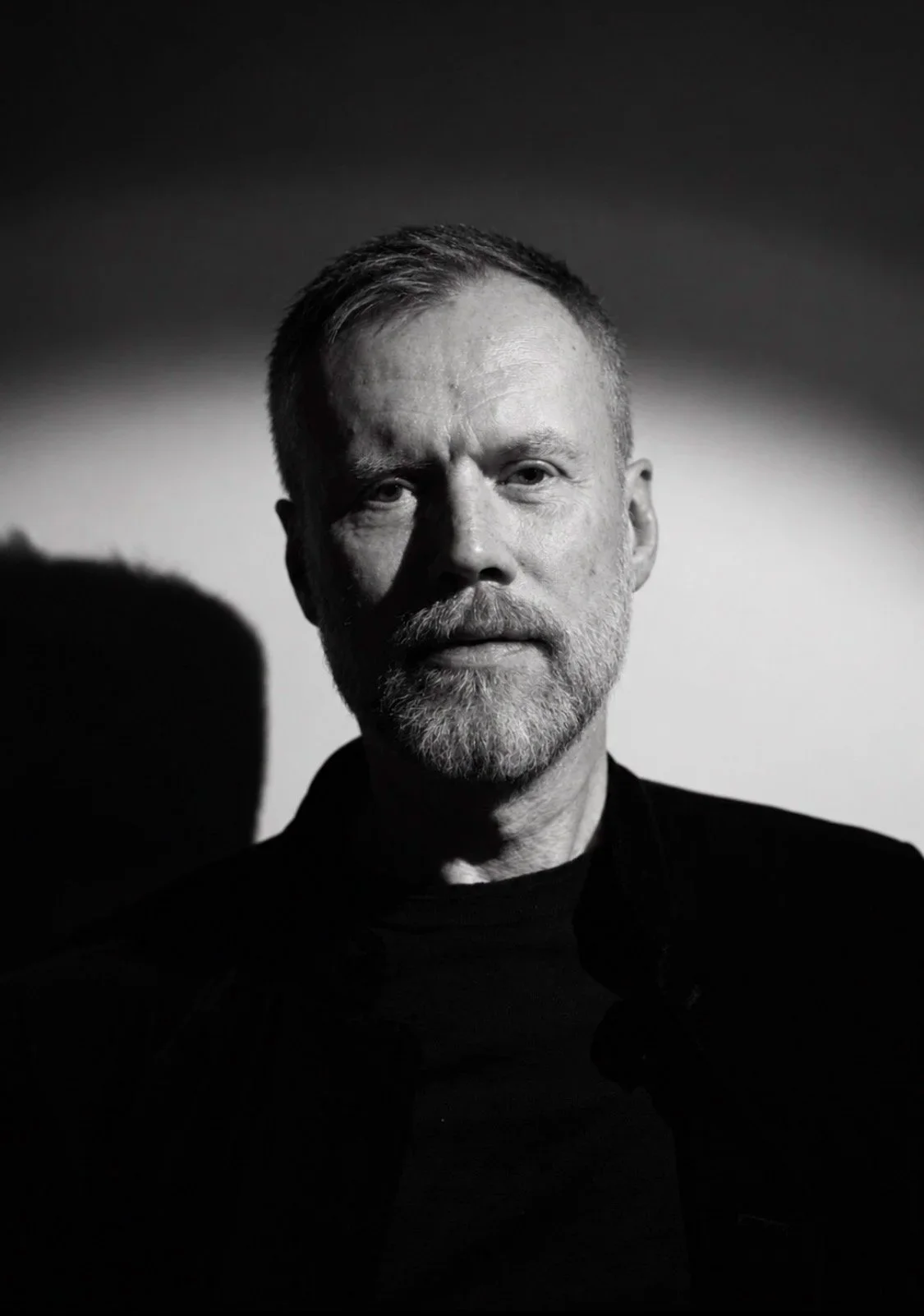 Black and white portrait of a middle-aged man with a beard and mustache, wearing a dark shirt, with light falling on one side of his face, creating shadows.