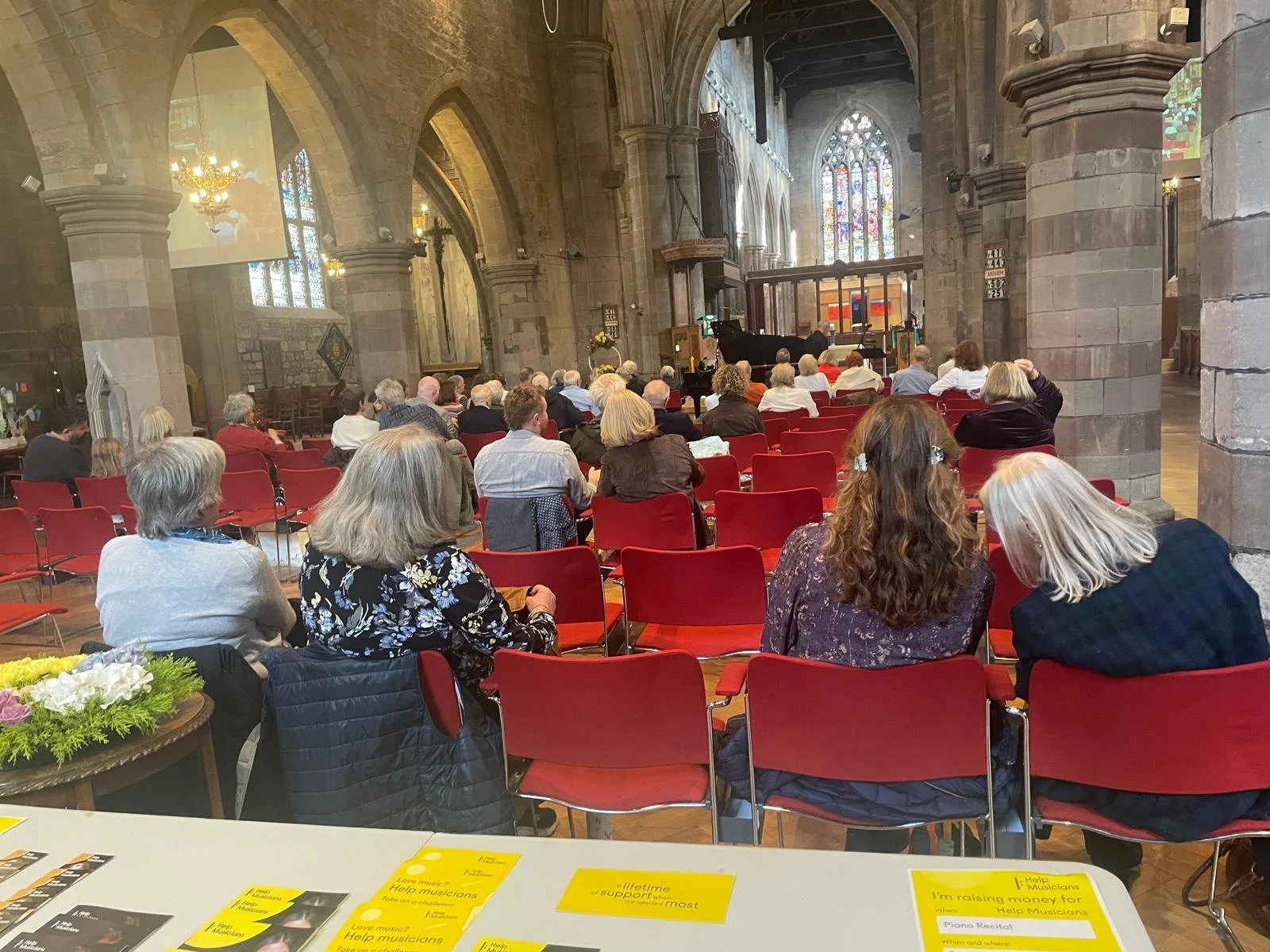 An audience seated in a church or cathedral with stone walls and stained glass windows, watching a concert with a pianist at a grand piano on stage.