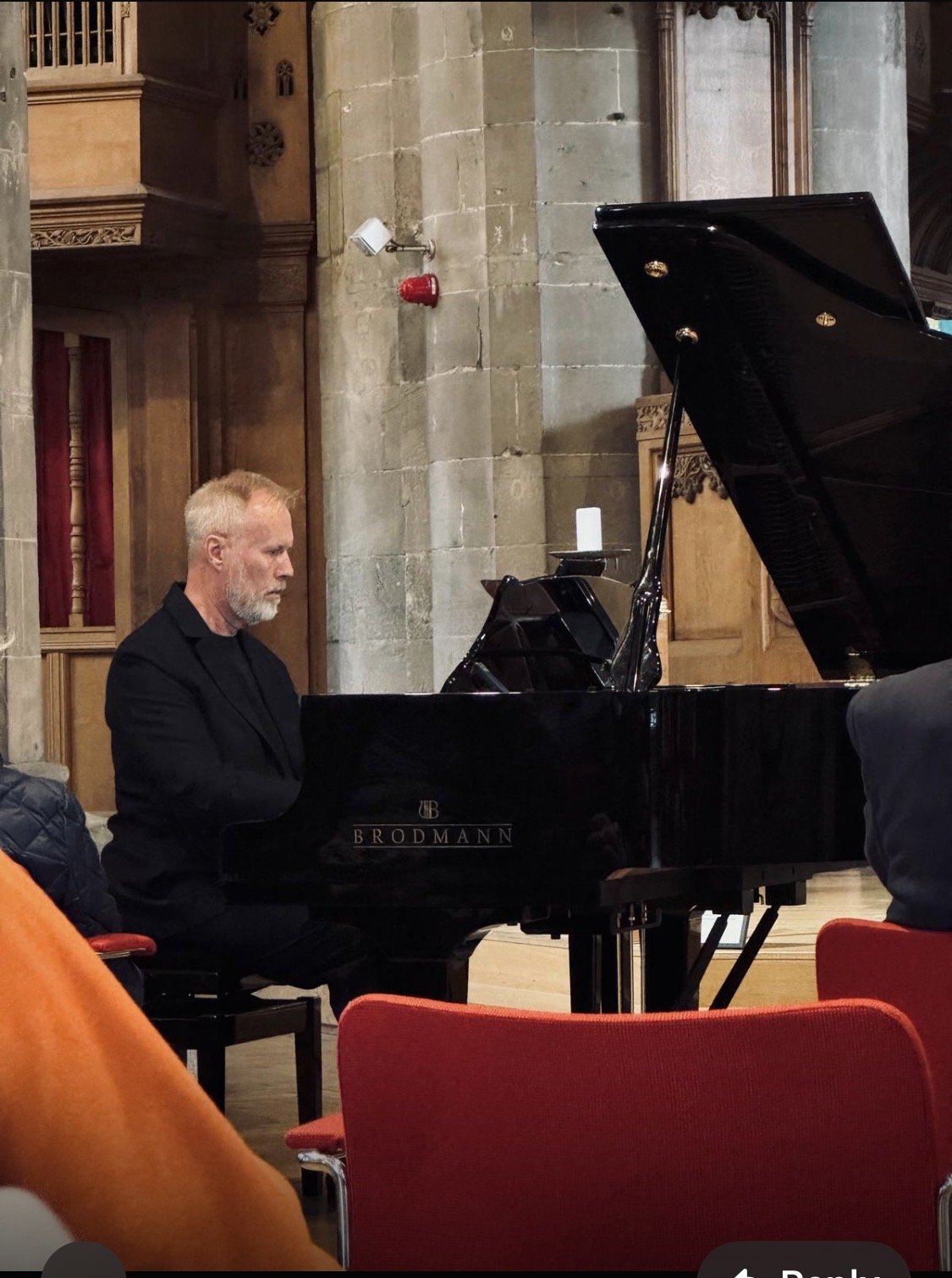 A man playing a black Brodmann grand piano in a church or cathedral with stone walls and wooden accents.