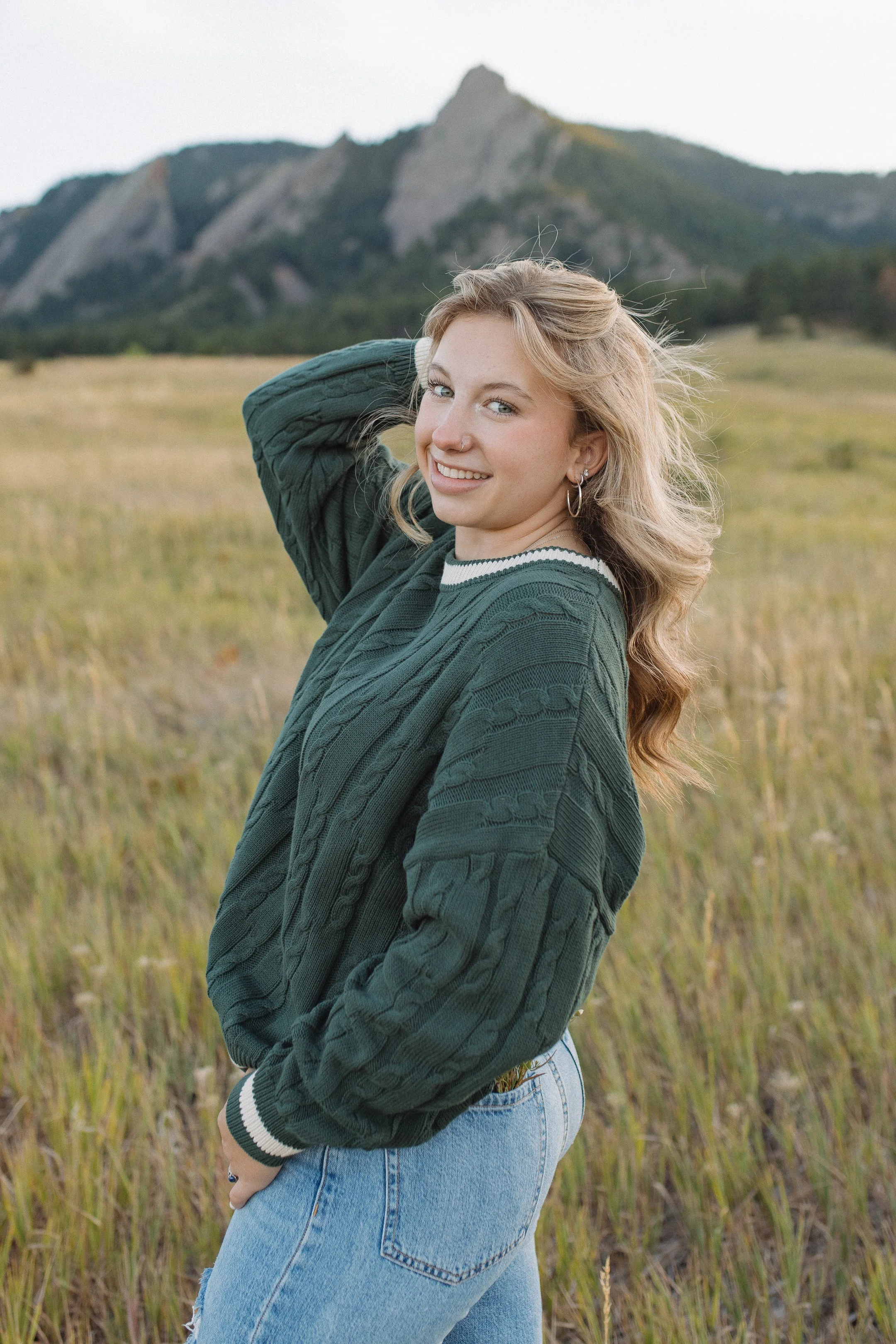 A young woman with wavy blonde hair smiling outdoors in a grassy field with mountains in the background, wearing a dark green sweater and blue jeans.
