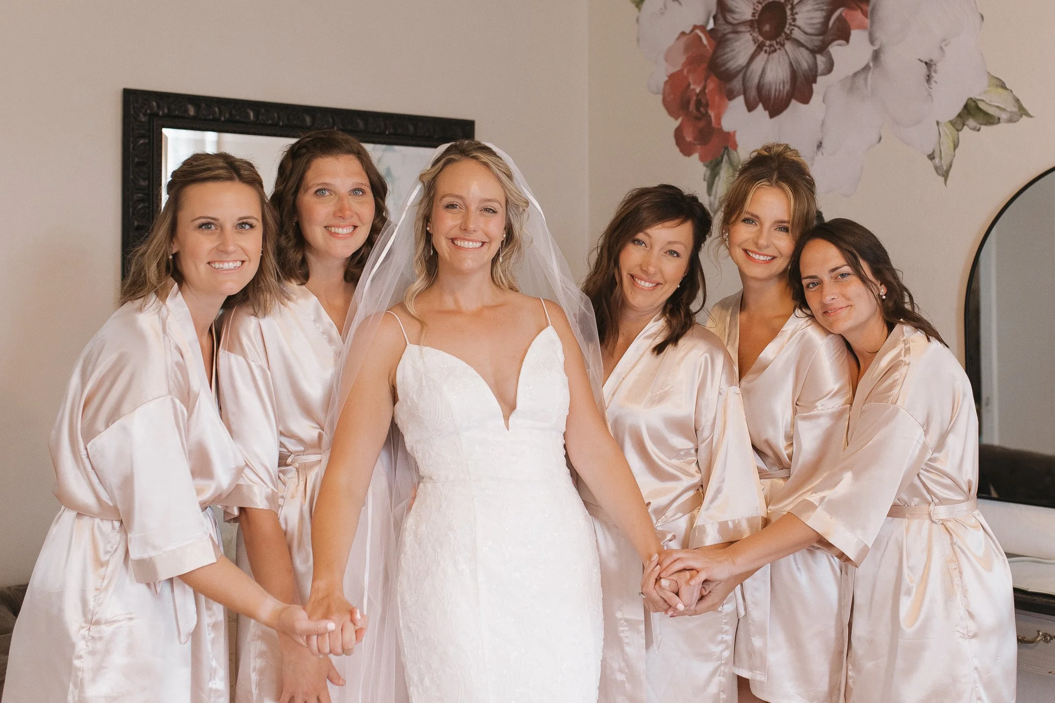 Bride surrounded by six women, all dressed in matching satin robes, smiling in a bridal suite with floral wall art and a mirror