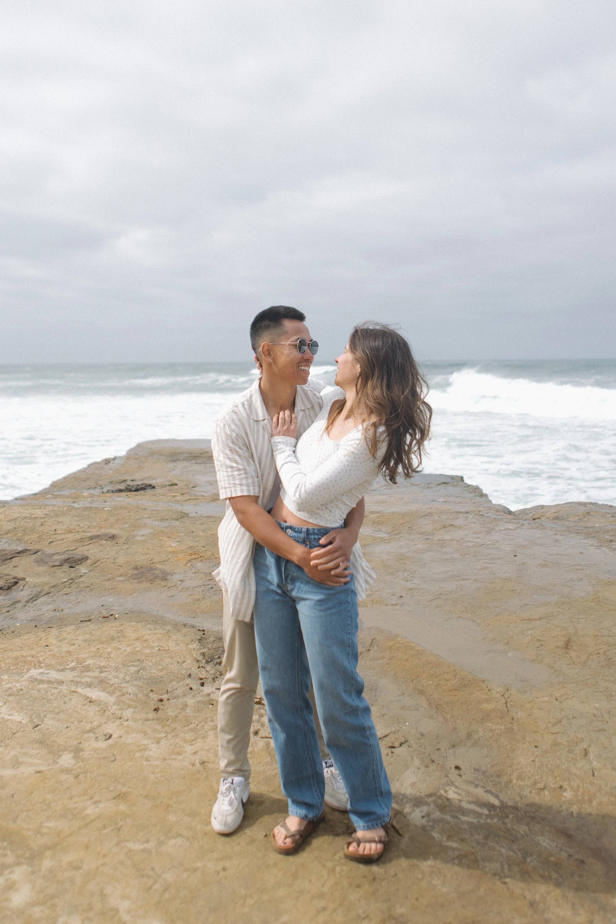 A young couple happily embraces on a rocky beach with ocean waves and a cloudy sky in the background.