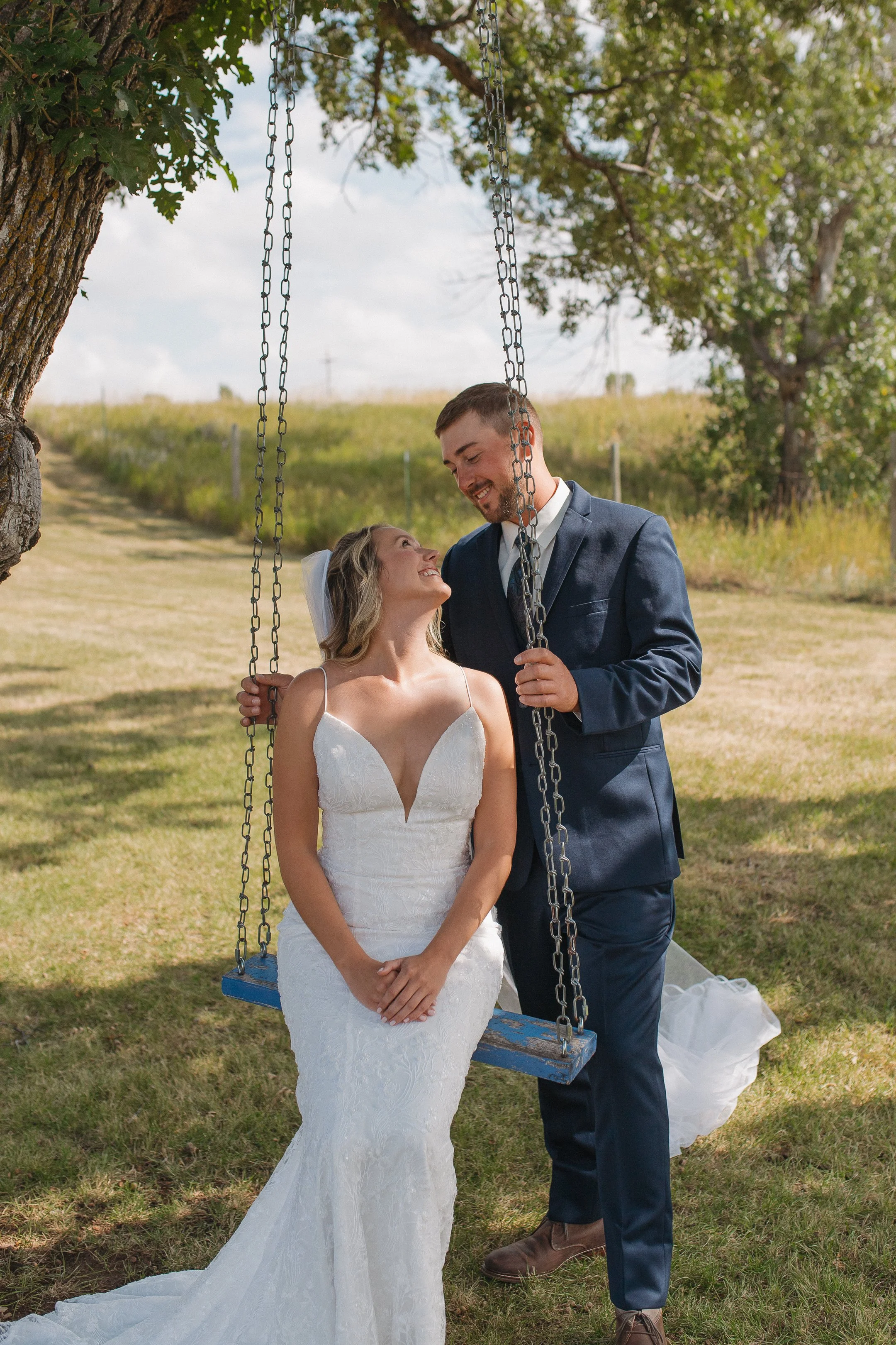 A bride and groom standing under a tree with a swing. The bride is sitting on the swing wearing a white wedding dress, and the groom, in a navy suit, is standing close to her, smiling and looking at her. It is an outdoor setting with green grass, tre