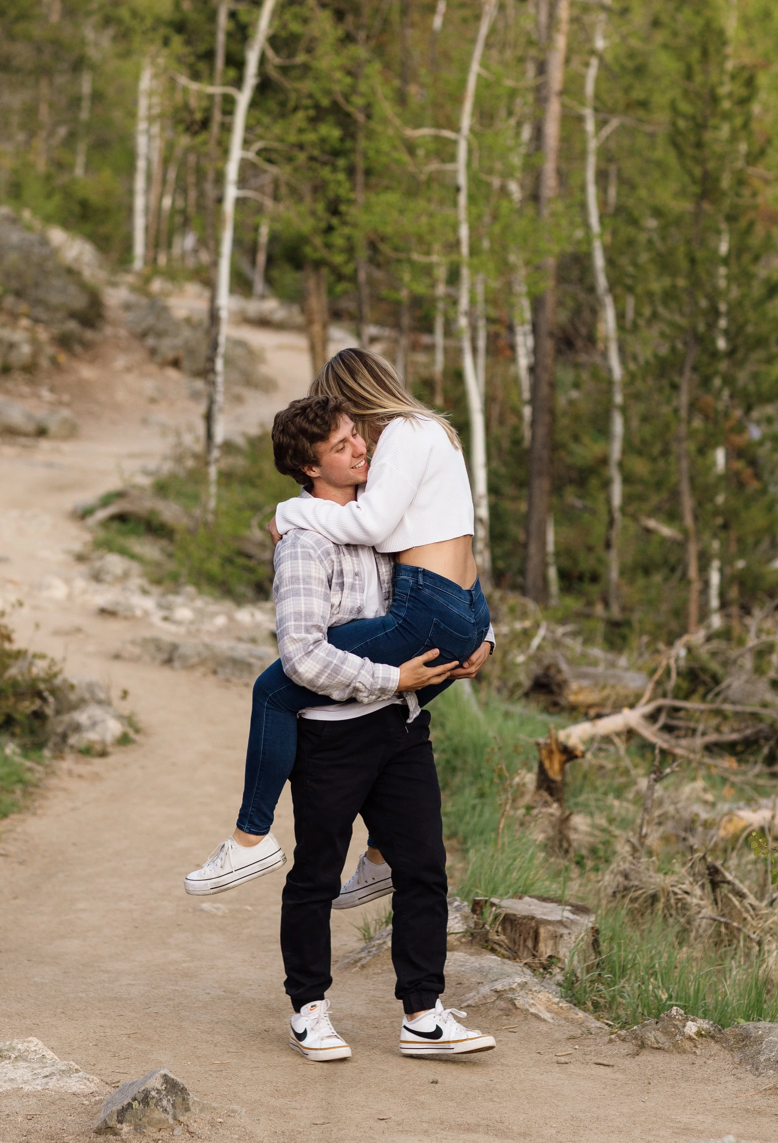A man carrying a woman in a forested area, both smiling and embracing.