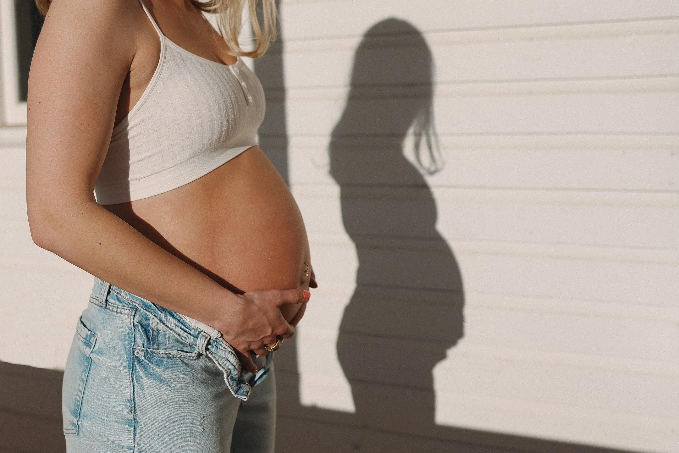 A pregnant woman wearing a white tank top and light blue denim shorts, standing outdoors in front of a white wall, with her shadow cast on the wall.