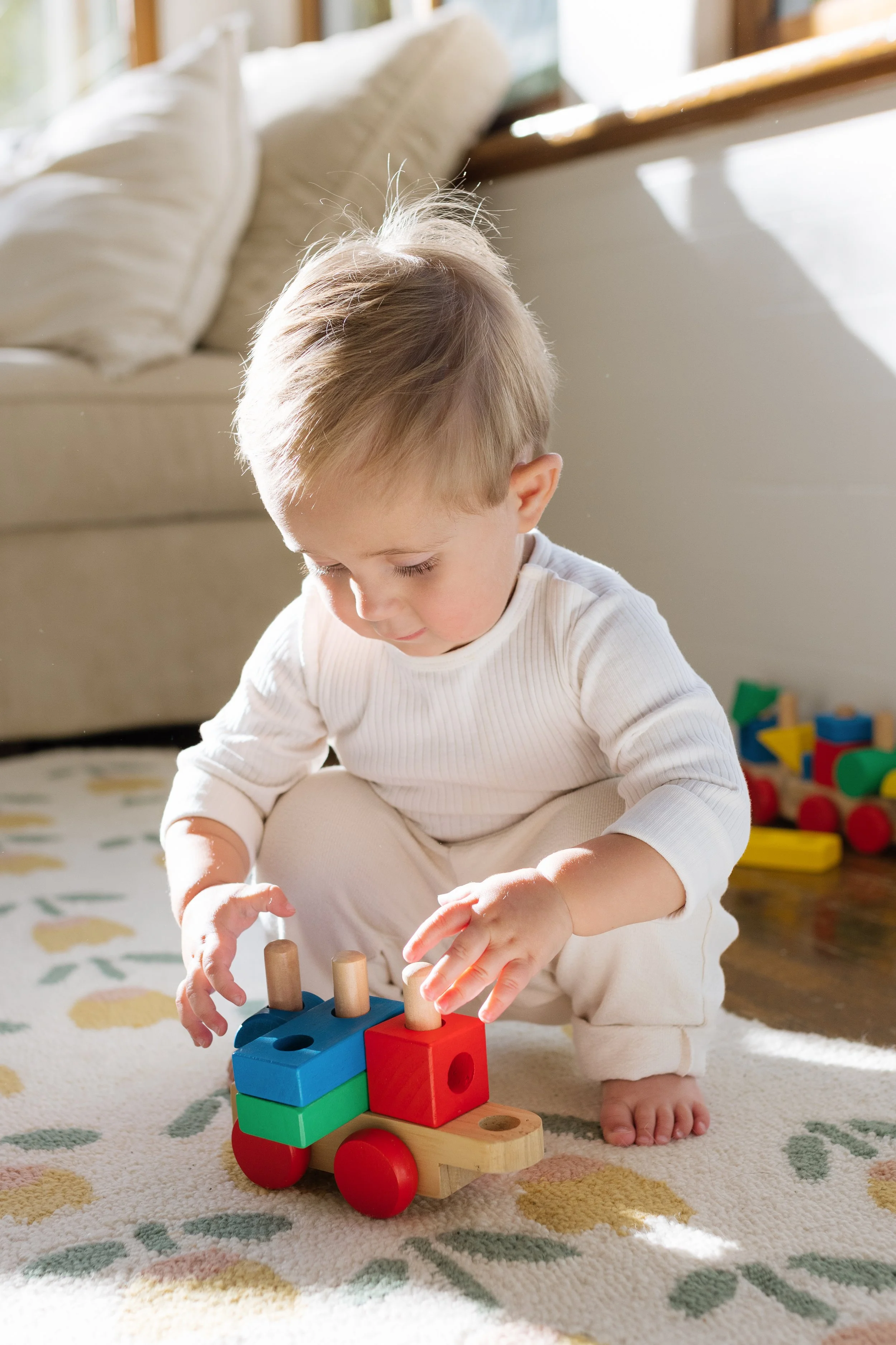 A young child playing with a colorful wooden toy train on a patterned rug in a sunlit room.