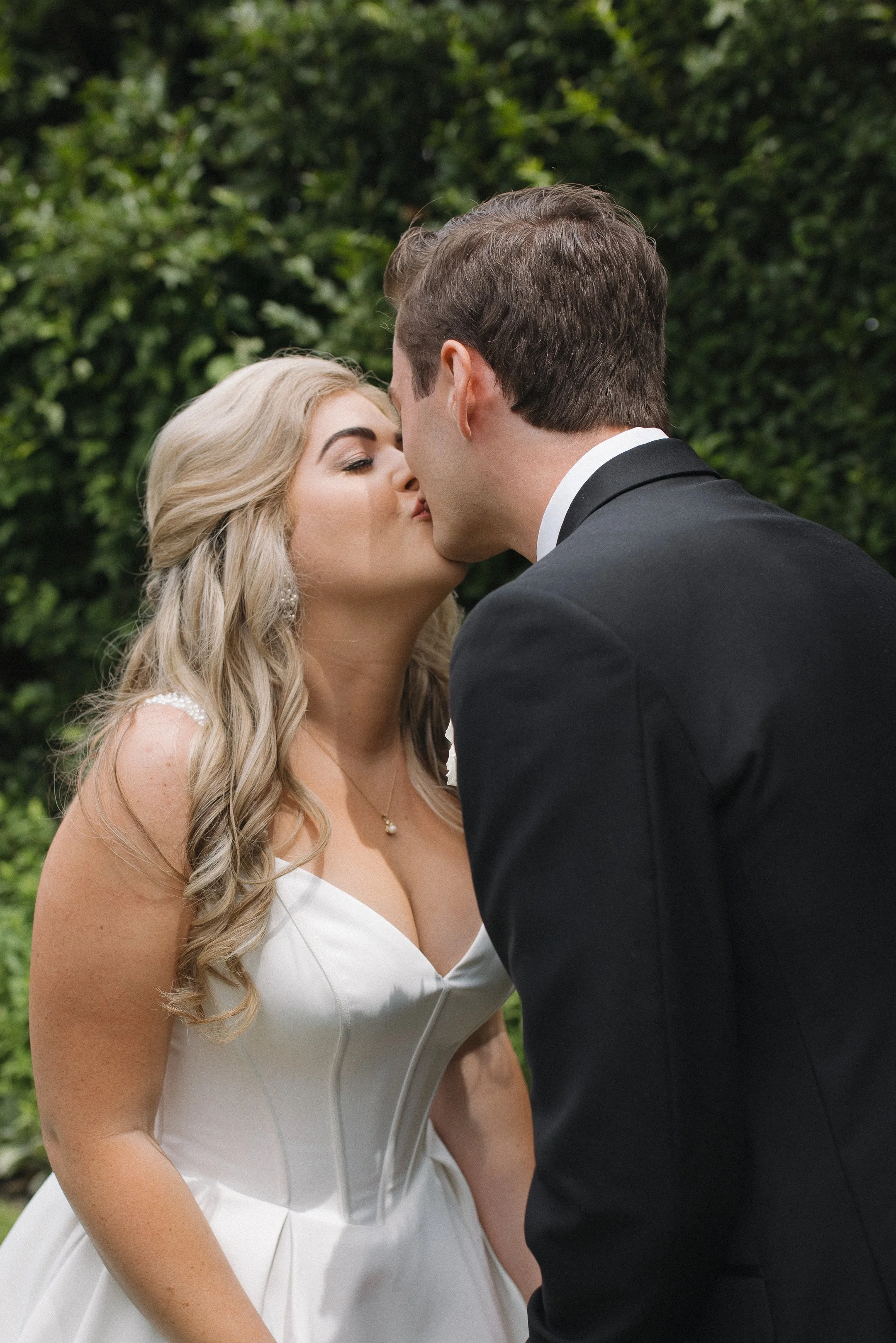 A bride and groom kiss outdoors, with the bride in a white wedding gown and the groom in a black suit, against a green foliage background.